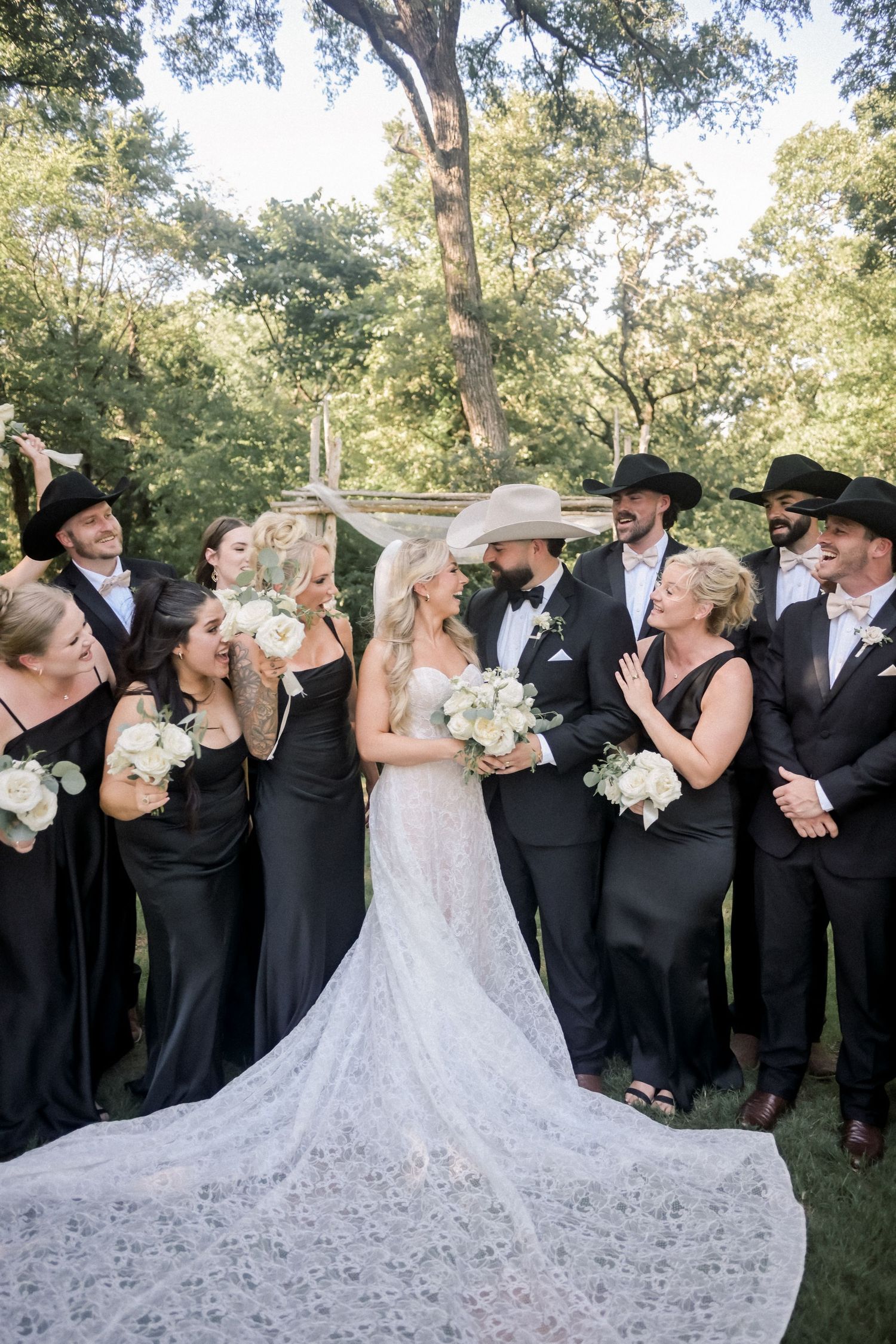 Wedding party in black attire and cowboy hats surrounds bride in lace gown and groom in tuxedo outdoors.
