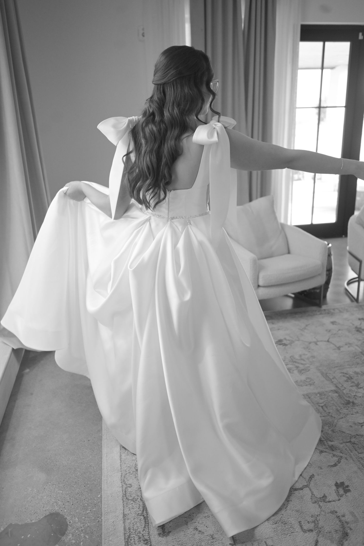 Bride in a stunning white satin gown with bow shoulders viewed from behind in a bright bridal suite.