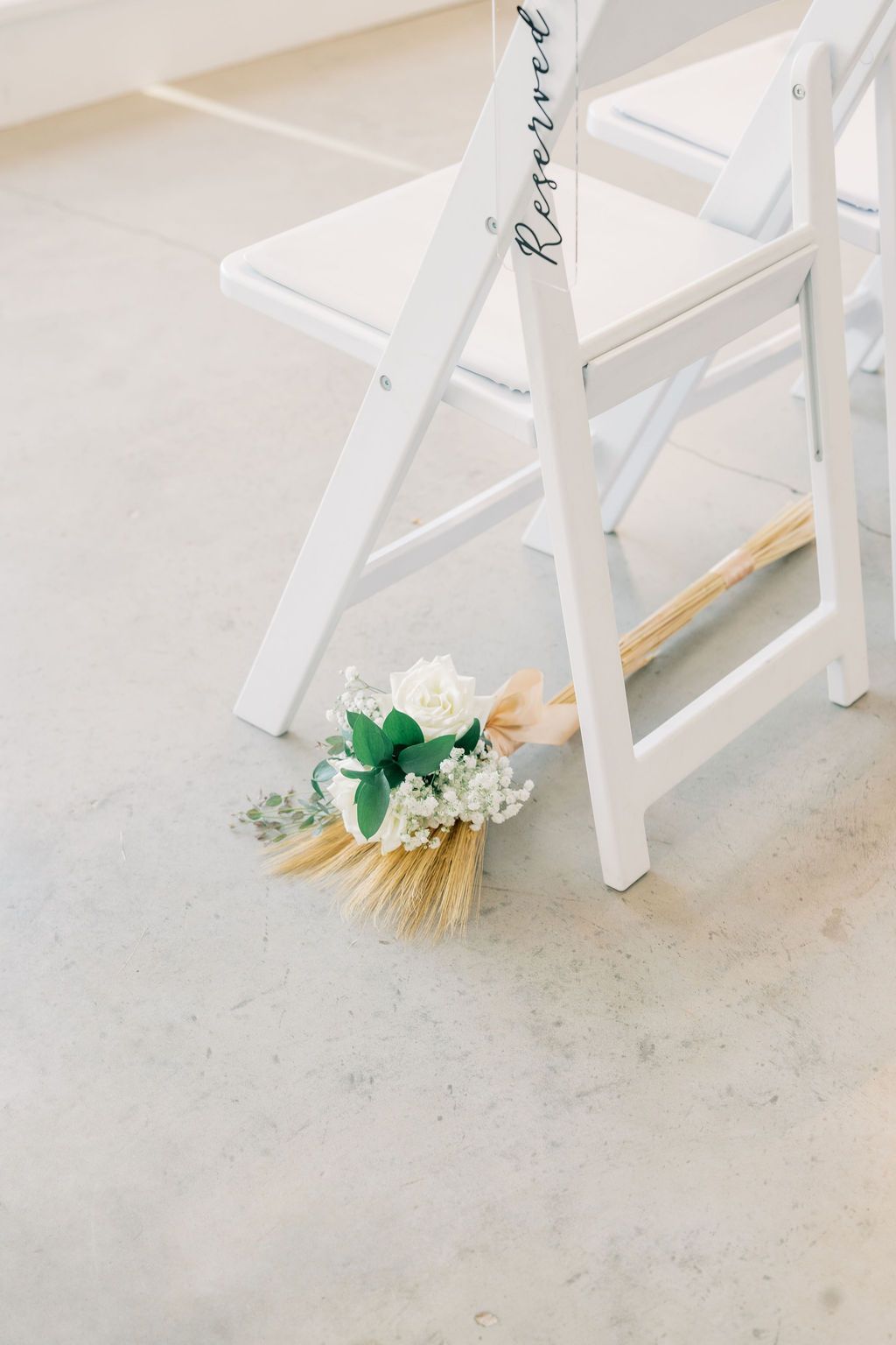 White folding chair with Reserved sign and small bridal bouquet of white flowers and wheat stems on concrete floor.