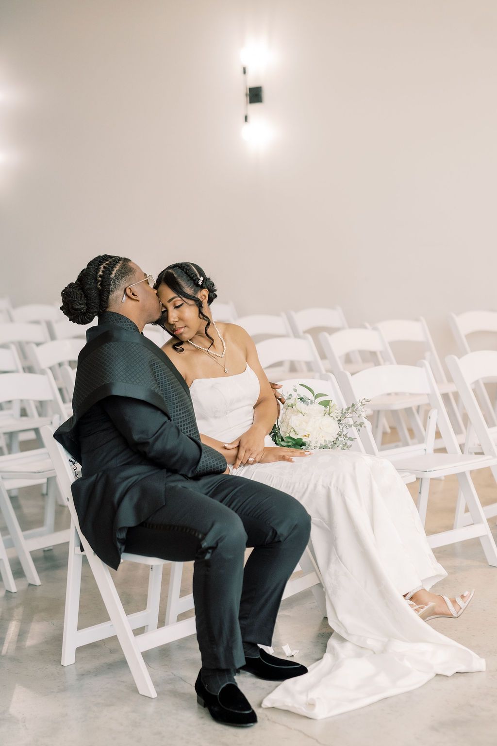 Bride and groom share a tender kiss while seated among white chairs in an elegant wedding venue.
