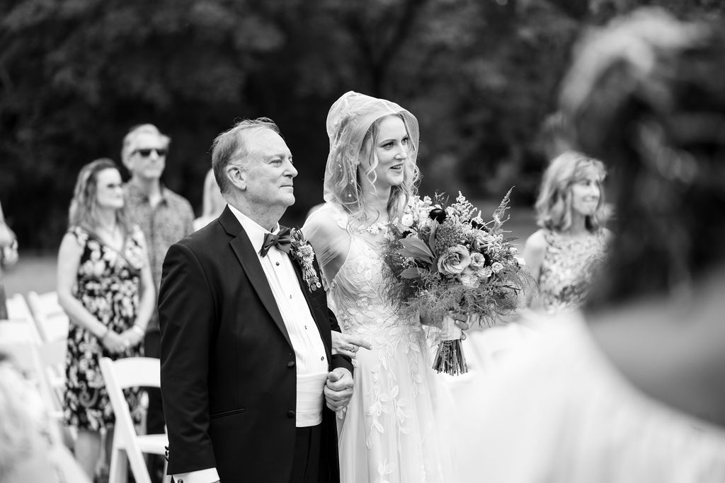 Black and white photo of a bride holding a bouquet walking down the aisle with an older man at an outdoor wedding.