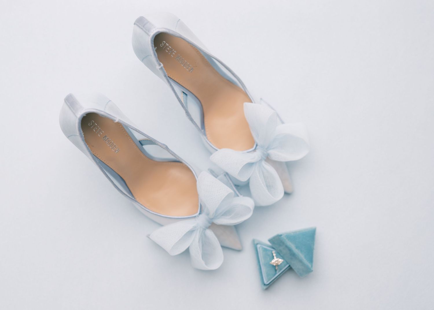 Light blue bridal heels with white bow accents beside a teal velvet ring box on a white background.
