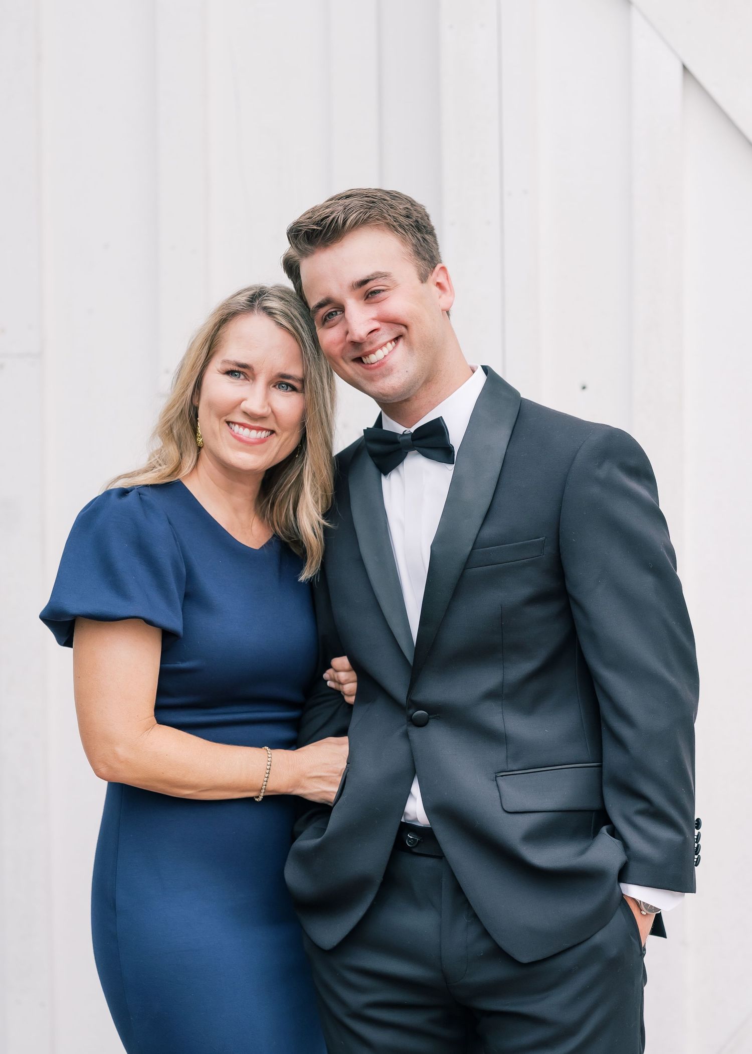 Smiling young man in dark tuxedo with bow tie posing with a woman in a navy blue dress.