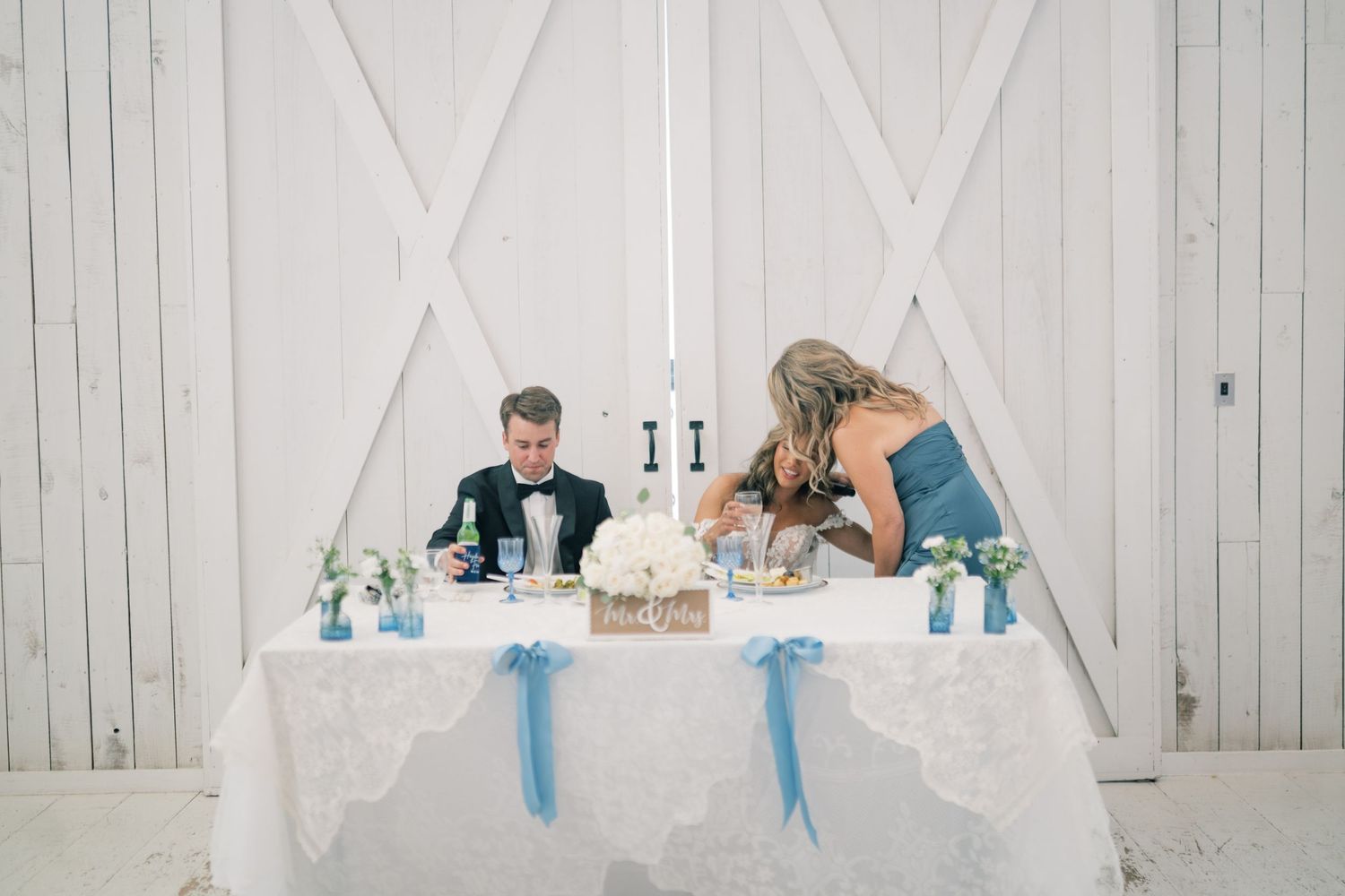 Bride and groom signing marriage documents at a lace-covered sweetheart table with blue ribbon accents.