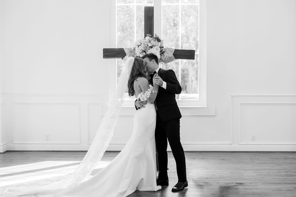 Bride and groom share first kiss before wooden cross in elegant white chapel wedding ceremony black and white photo.