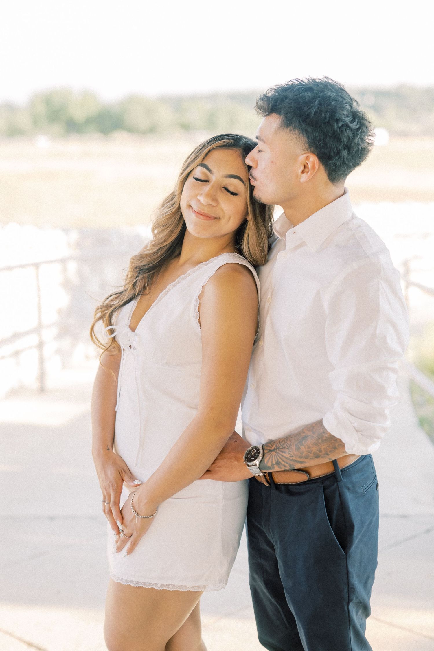 Man in white shirt kissing woman in white mini dress during outdoor engagement photo session.