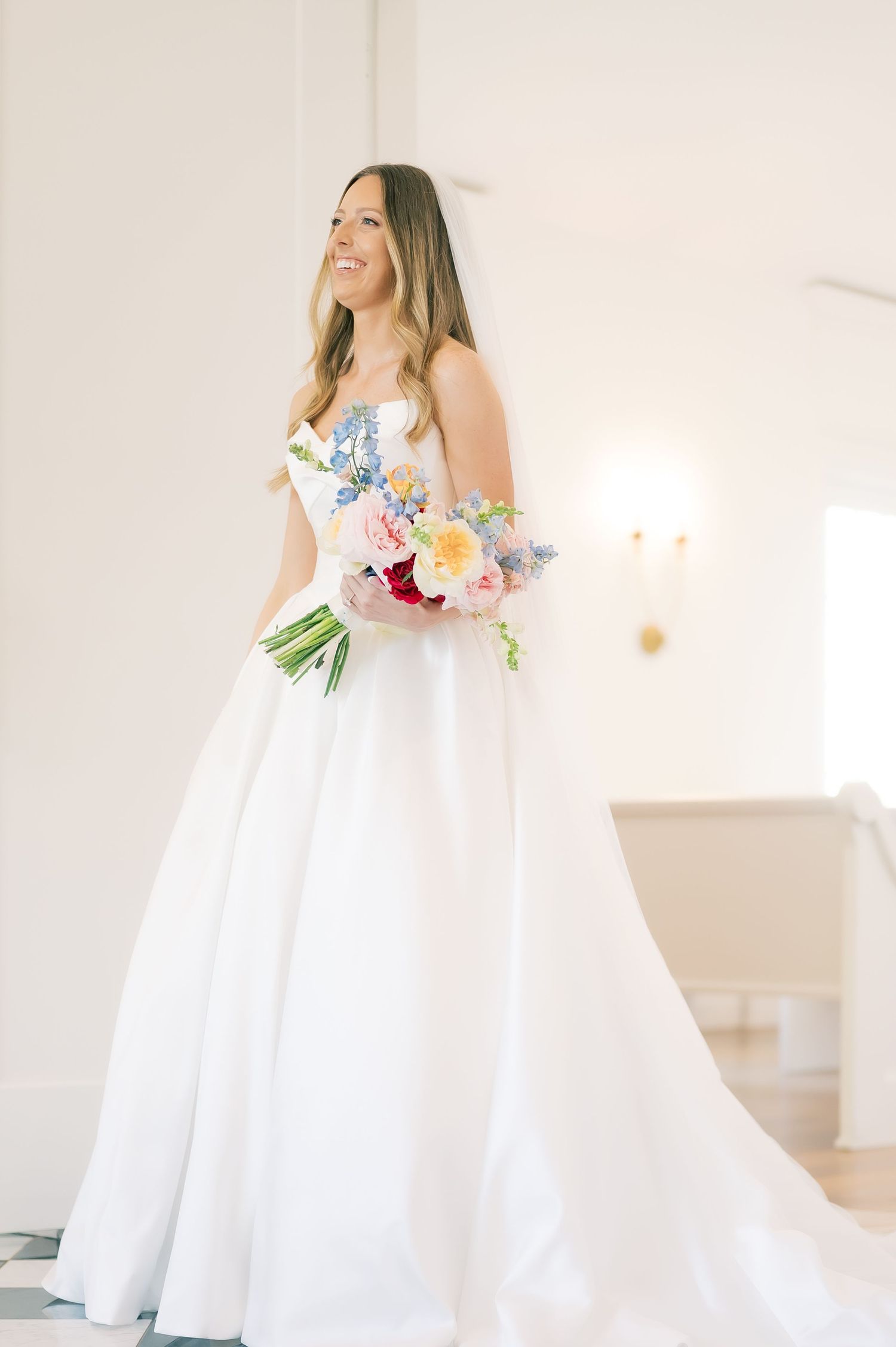 Smiling bride in strapless white ball gown holding colorful bouquet with pink, blue, and yellow flowers in bright chapel.