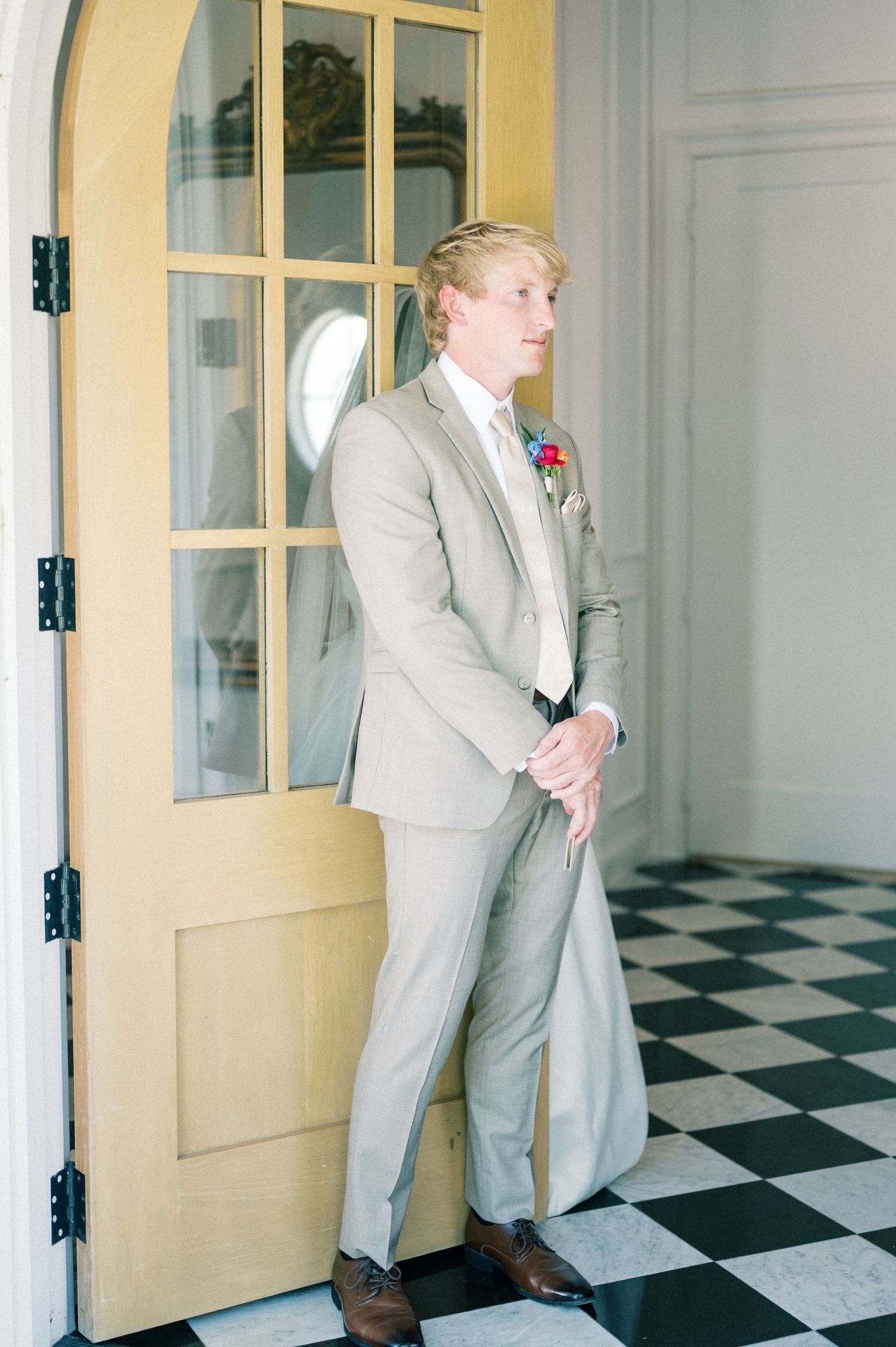Groom in tan suit with boutonniere leaning against wooden door on black and white checkered floor.