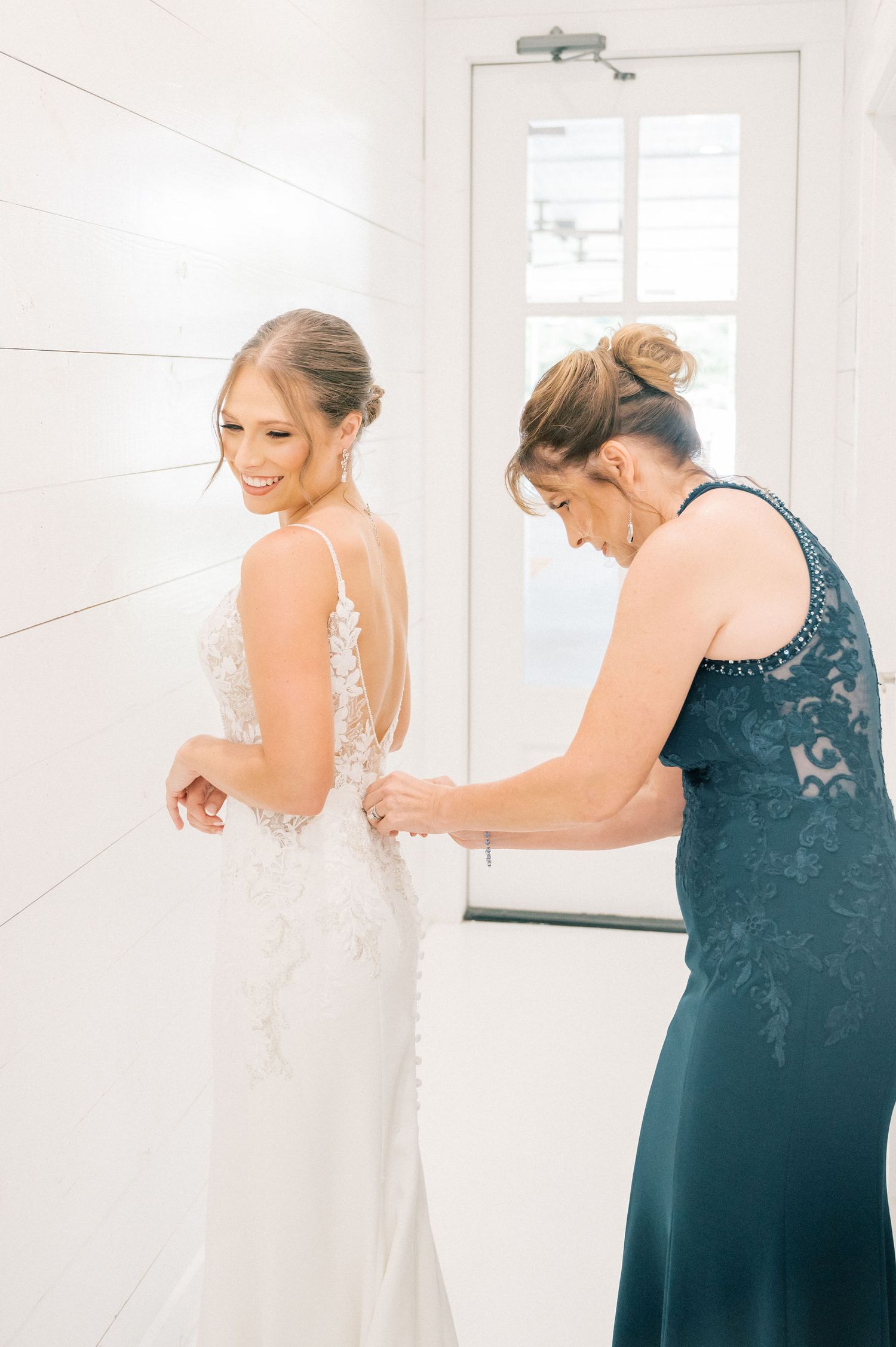 A person in a white wedding gown gets help with final dress preparations in a sunlit room.