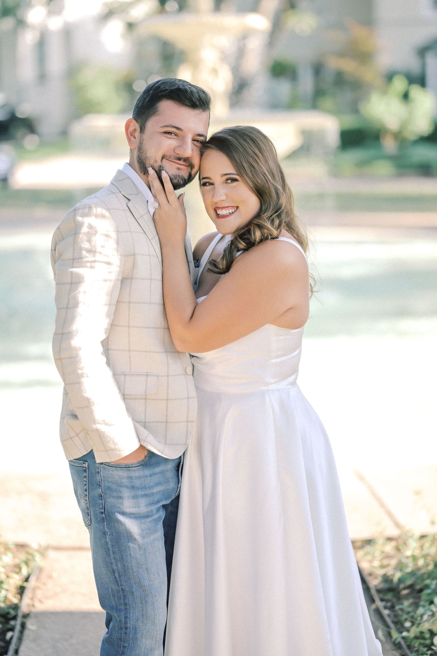 Smiling couple embracing outdoors in a sunlit park, he in a plaid blazer and jeans, she in a flowing white satin dress.