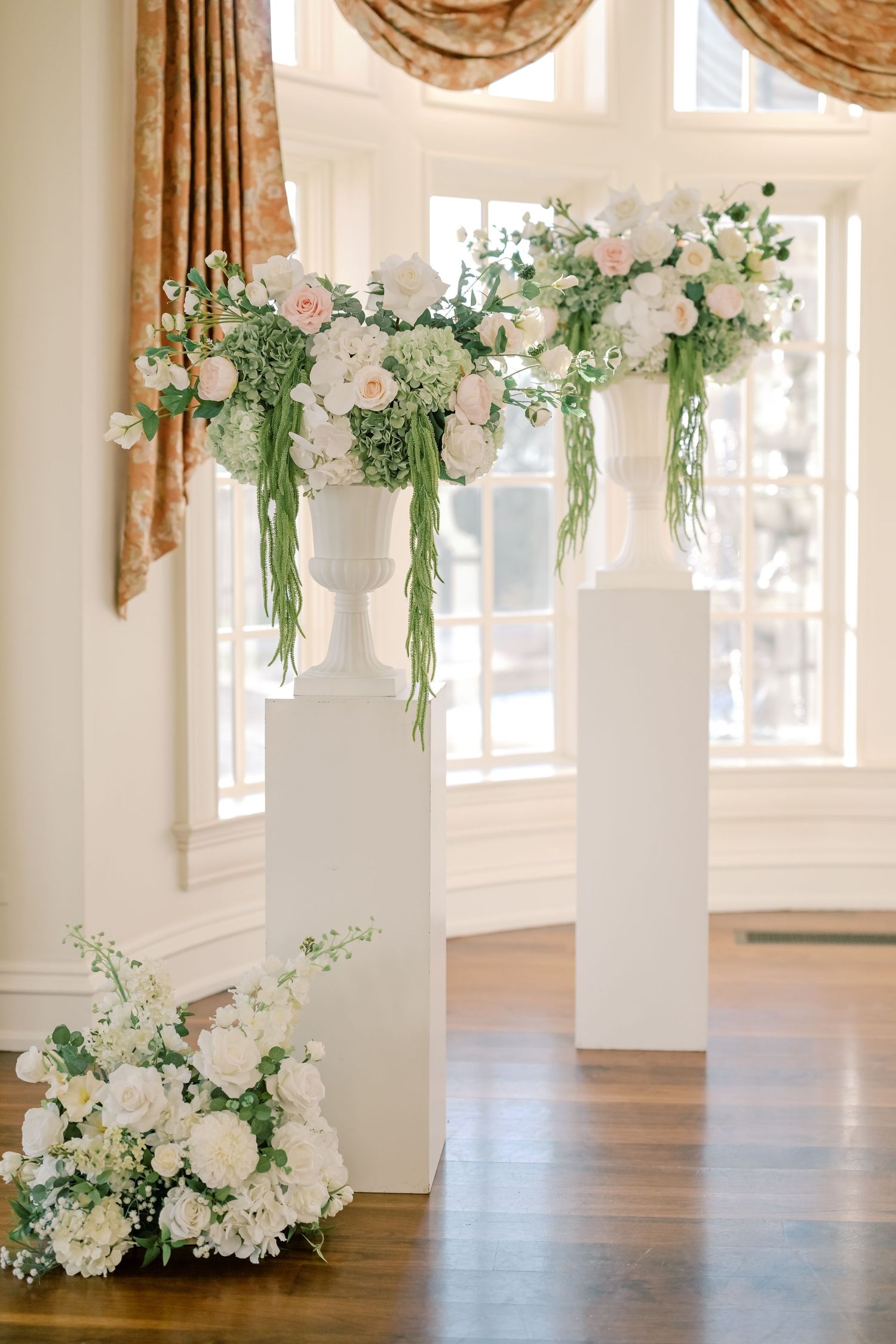 Elegant white floral arrangements on tall pedestals with cascading greenery decorate a bright wedding venue interior.