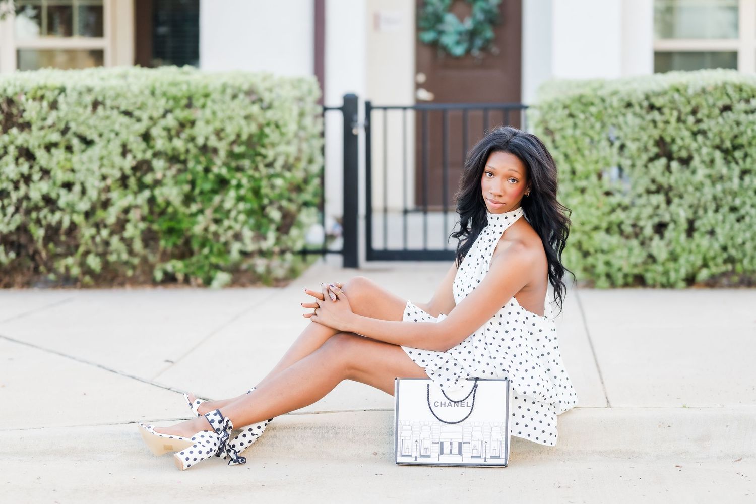 A young Black woman in a white polka dot dress sits gracefully on pavement beside a shopping bag outdoors. Seymour TX HS Senior Photography