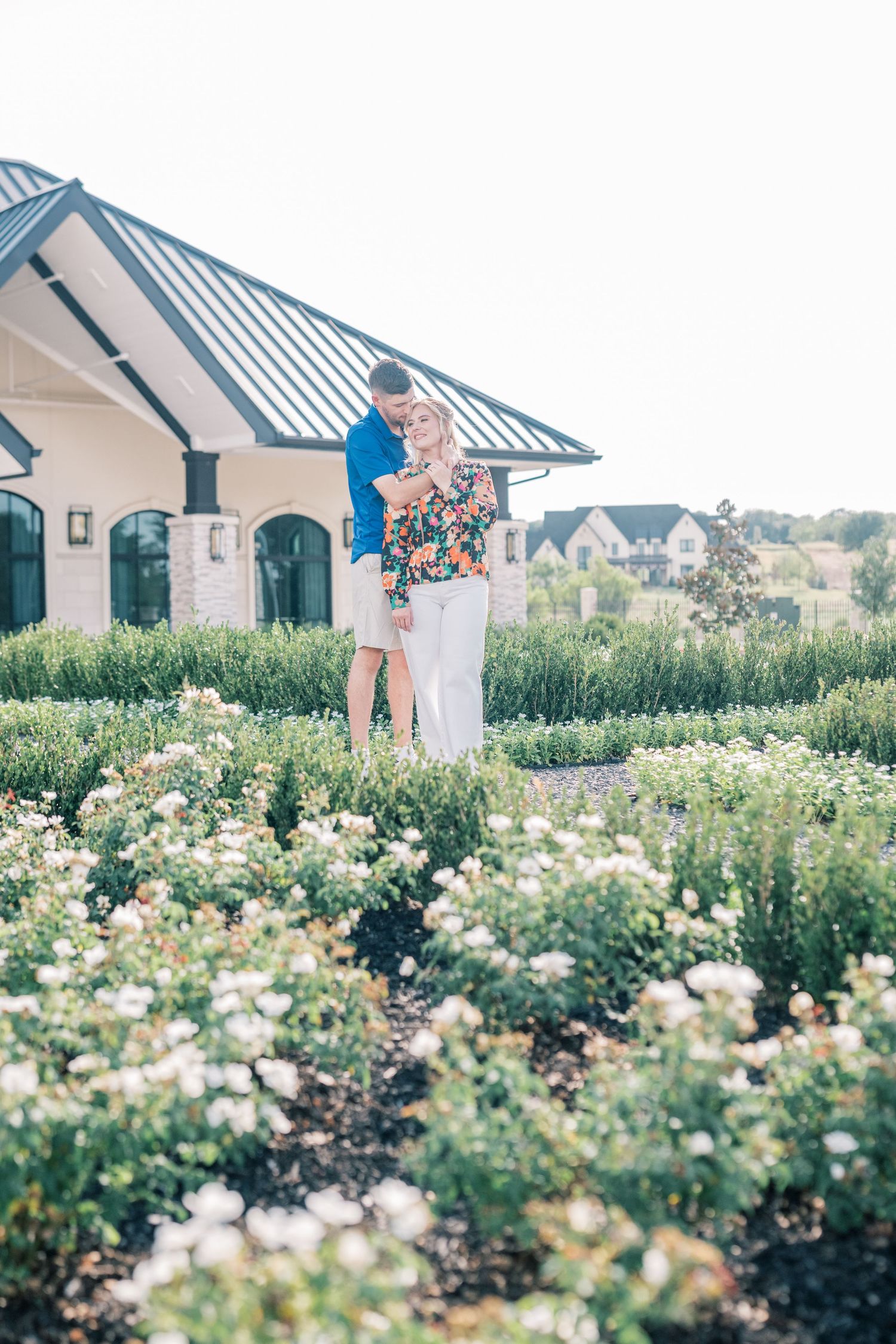 A couple embraces in a lush garden with white flowers outside an elegant white building.