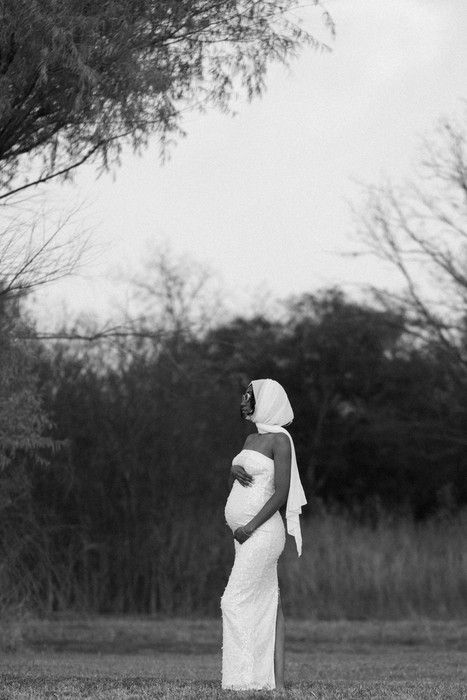 A figure in white stands contemplatively in a grassy field with trees in the background, captured in black and white.