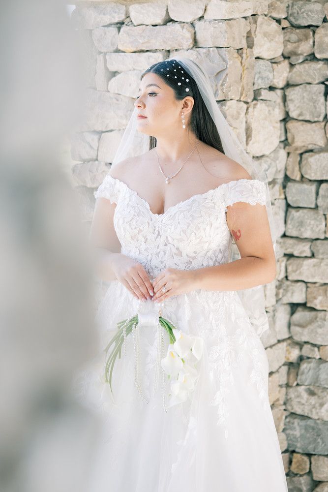 Ethereal bridal portrait against stone wall wearing off-shoulder lace gown and crystal headpiece.