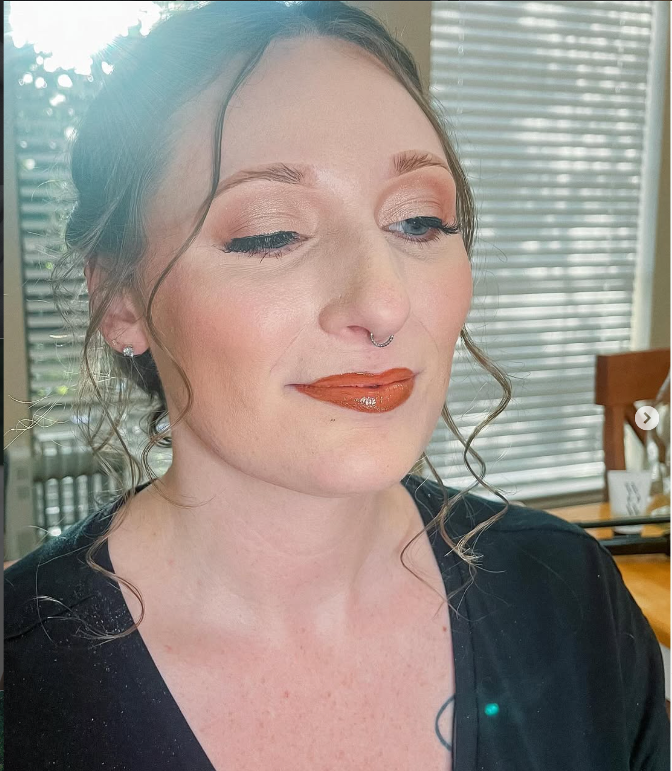 Woman with elegant updo, bold terracotta lips, and subtle eye makeup wearing a black top near a window.