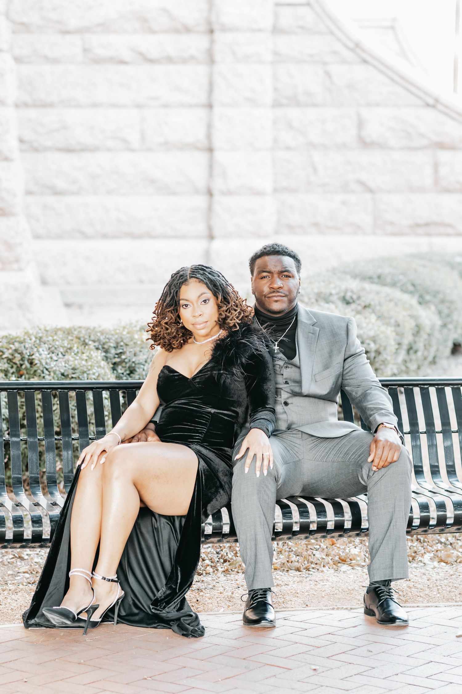 Stylish couple in black velvet gown and gray suit posing on a bench outdoors.