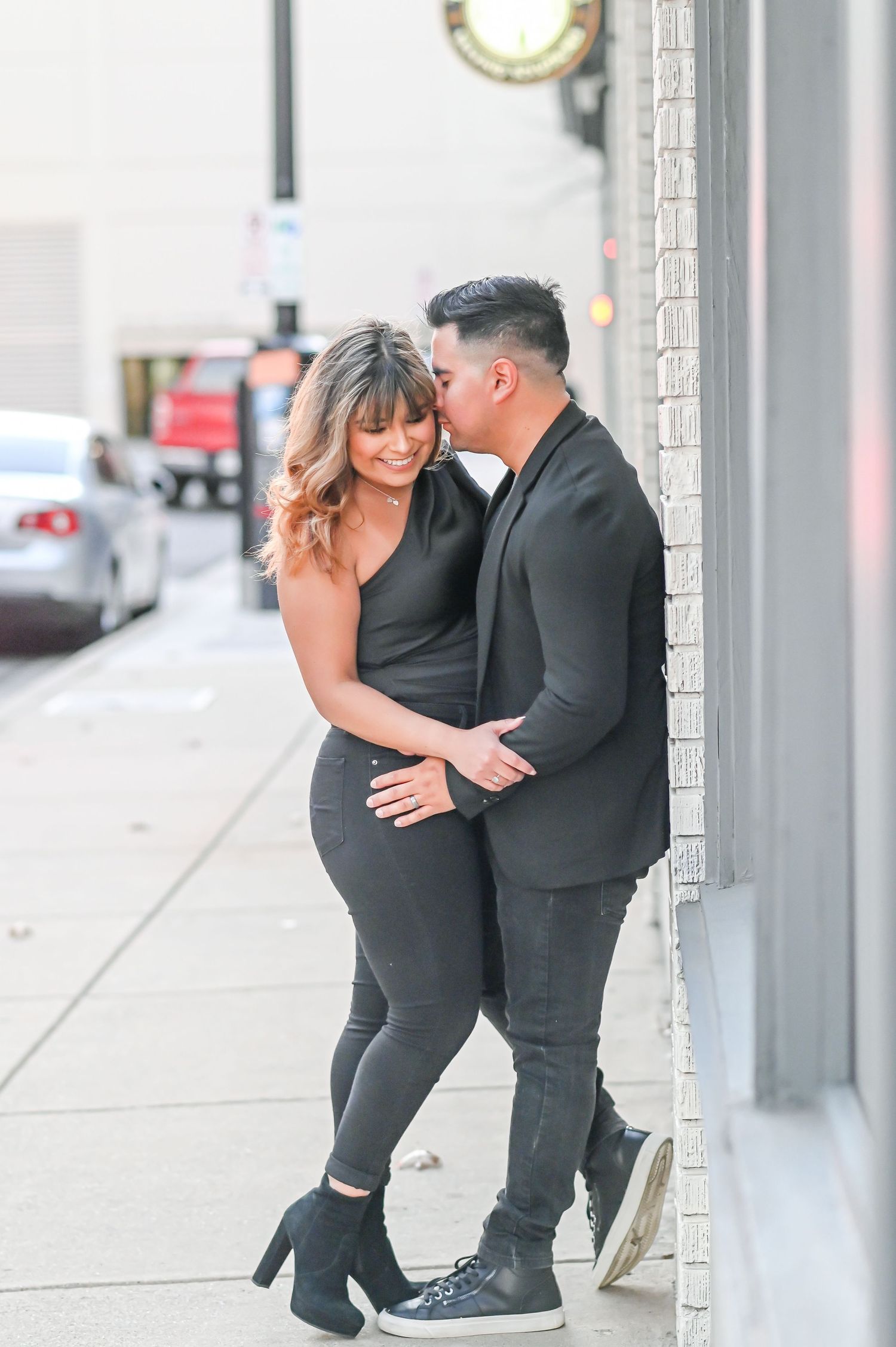 Couple in matching black outfits sharing a romantic moment on a city sidewalk, man kissing woman's cheek.