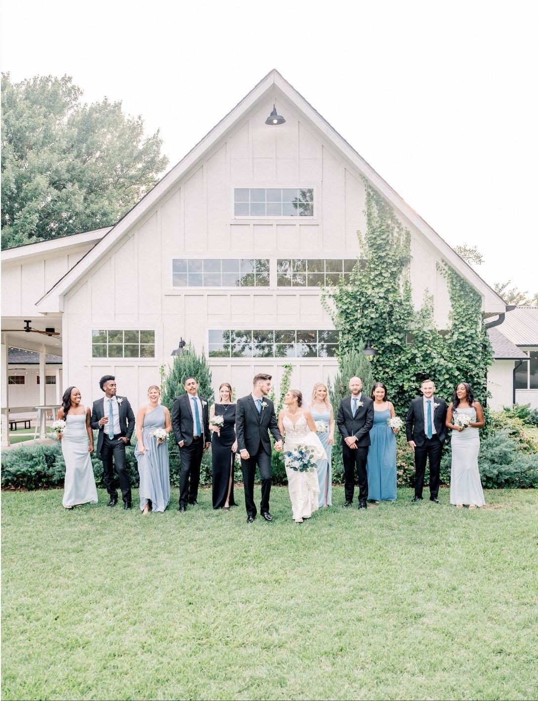 Wedding party posing in front of a white barn venue on a sunny day, bridesmaids in blue gowns and groomsmen in black suits.