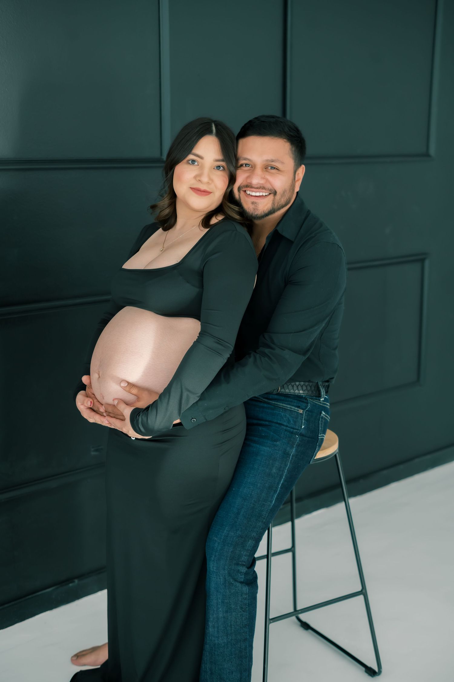 Smiling couple in dark green outfits pose together during maternity photoshoot, man embracing pregnant woman from behind.