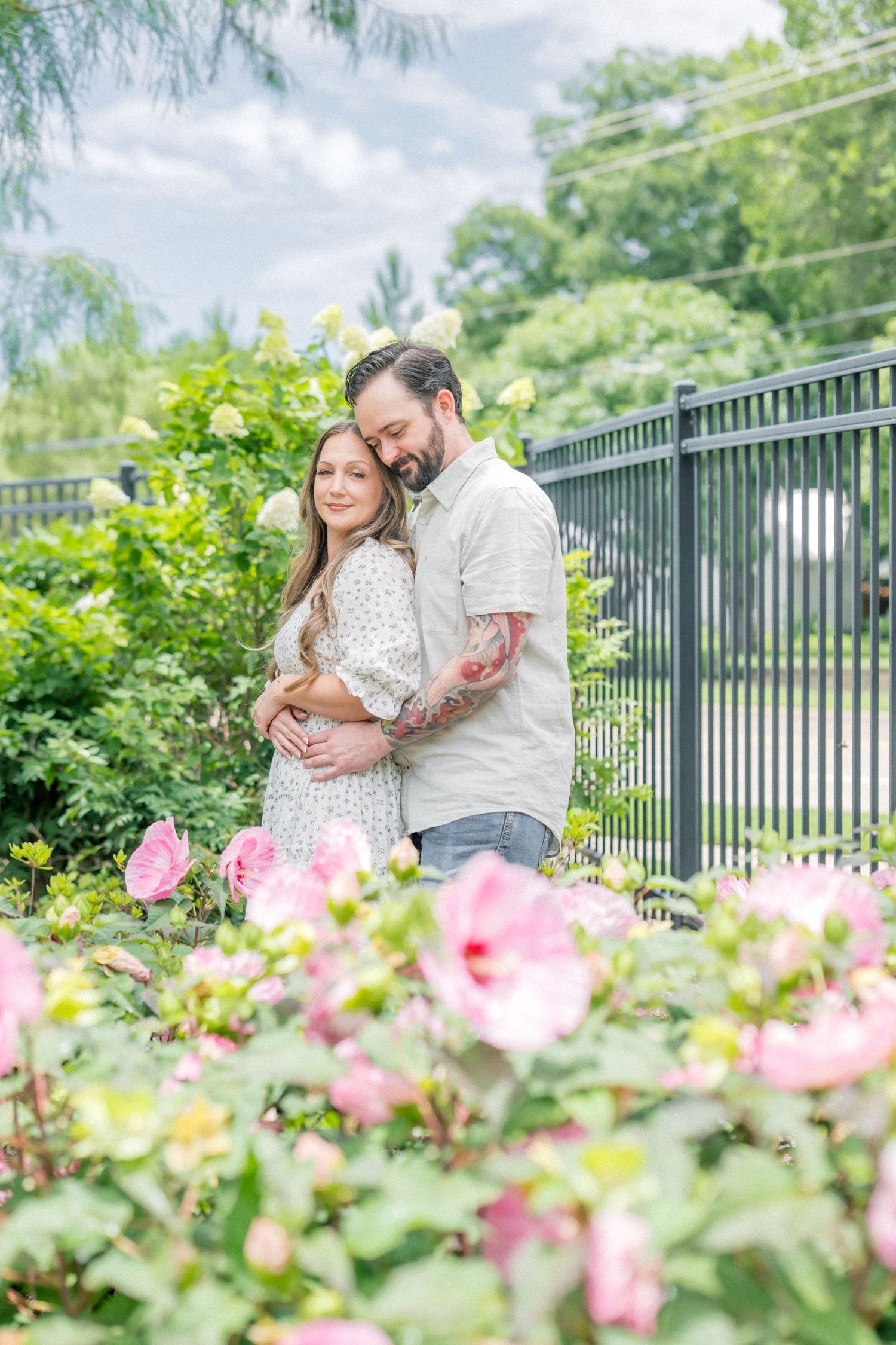 Couple embracing tenderly among pink roses and greenery near a black iron fence in a garden.
