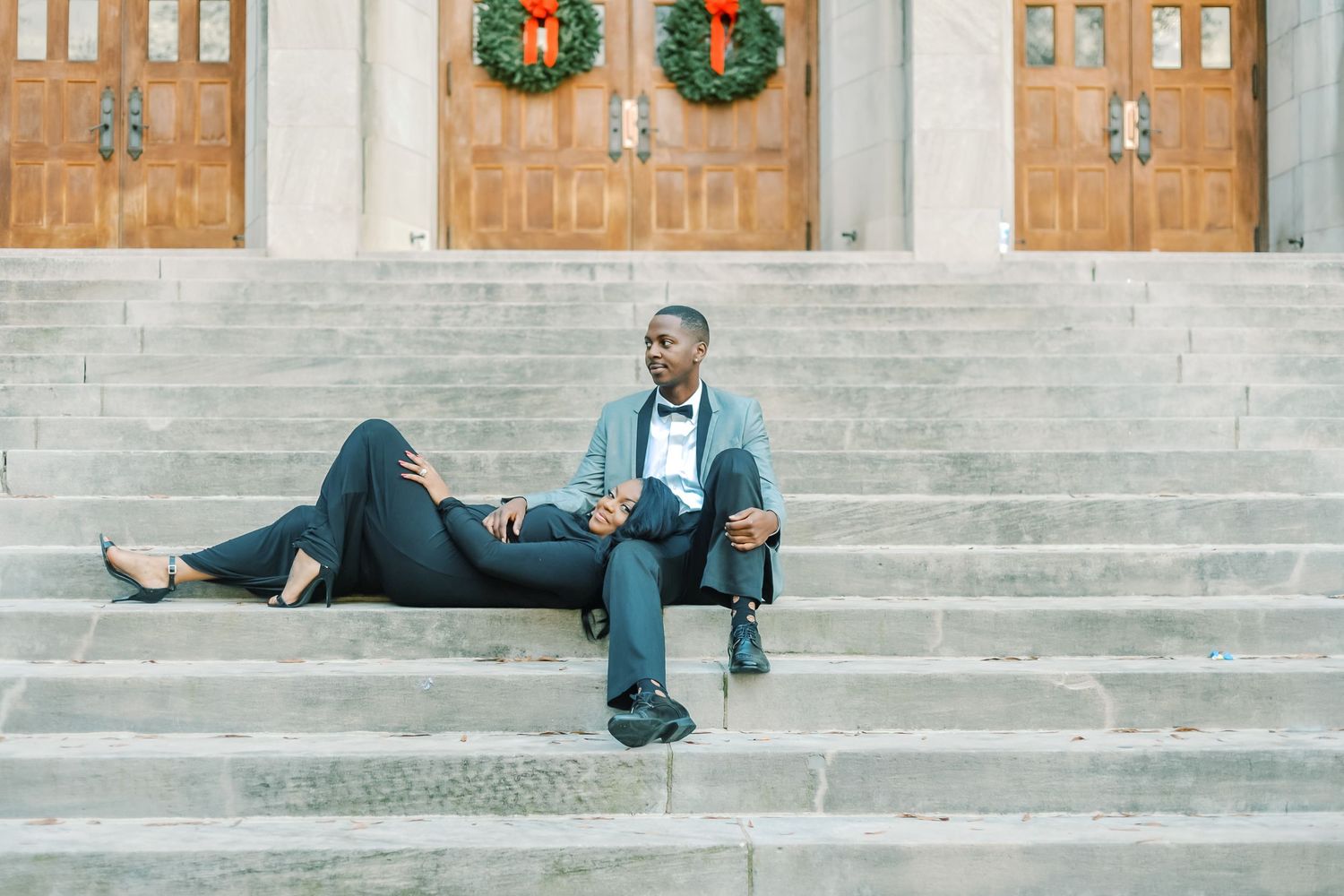Stylish couple relaxing on stone church steps with Christmas wreaths decorating the doors behind them.