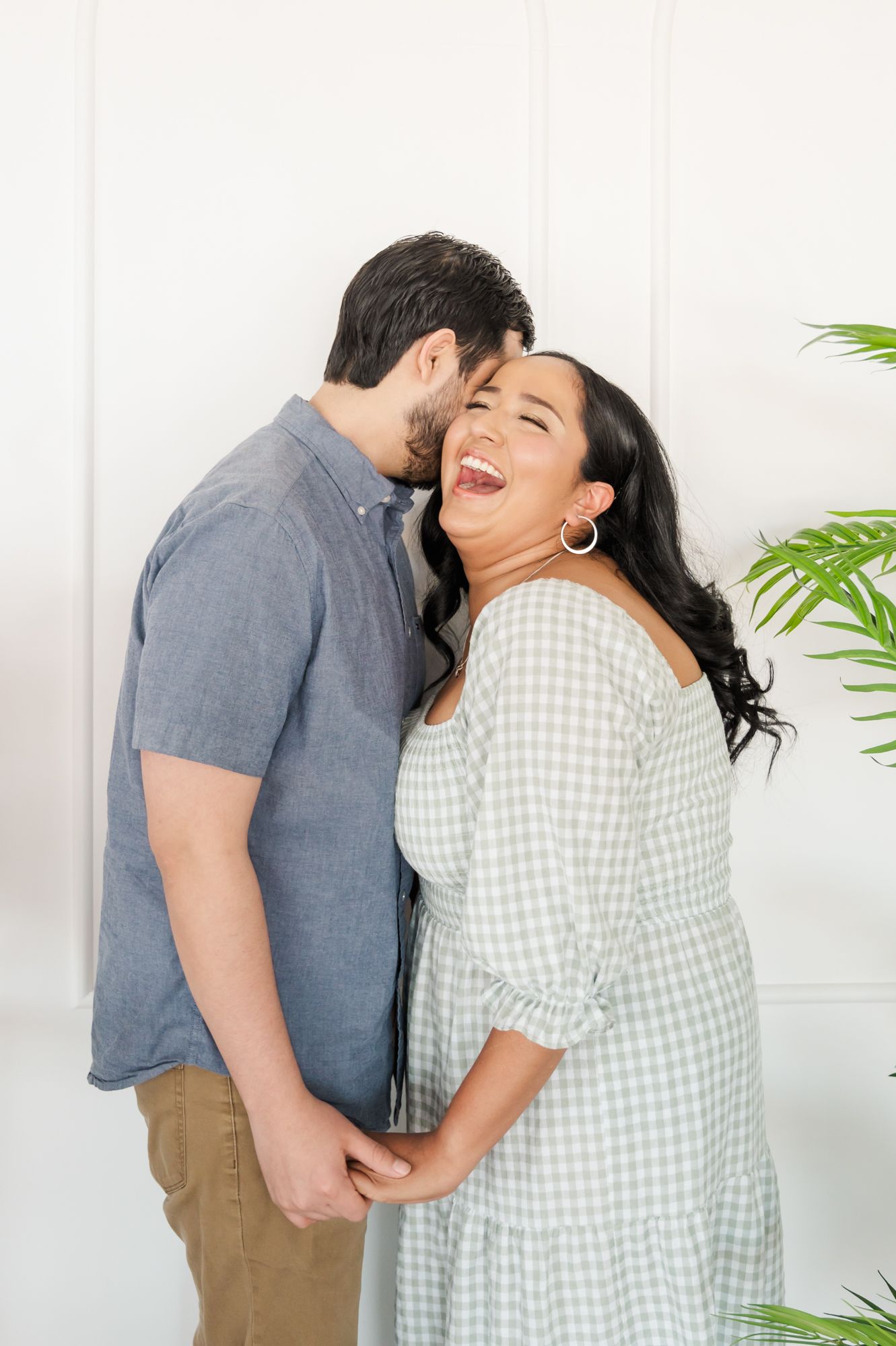A joyful couple sharing an intimate moment against a white wall with green plants in the background.