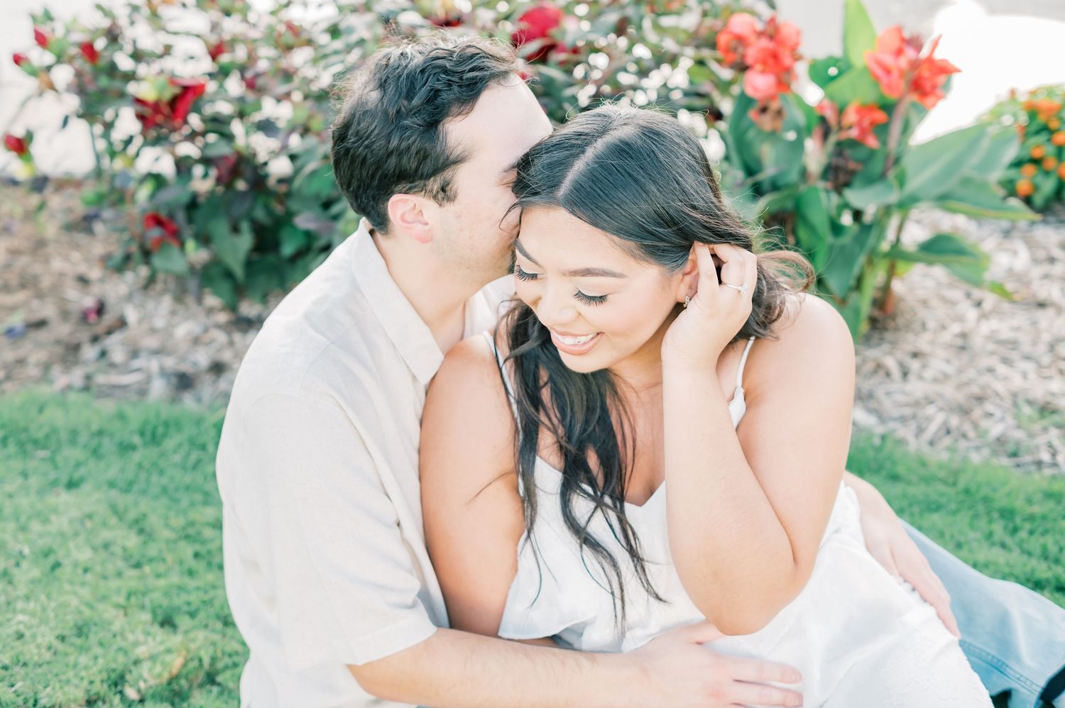 A couple shares an intimate moment in a garden setting with vibrant red flowers in the background.