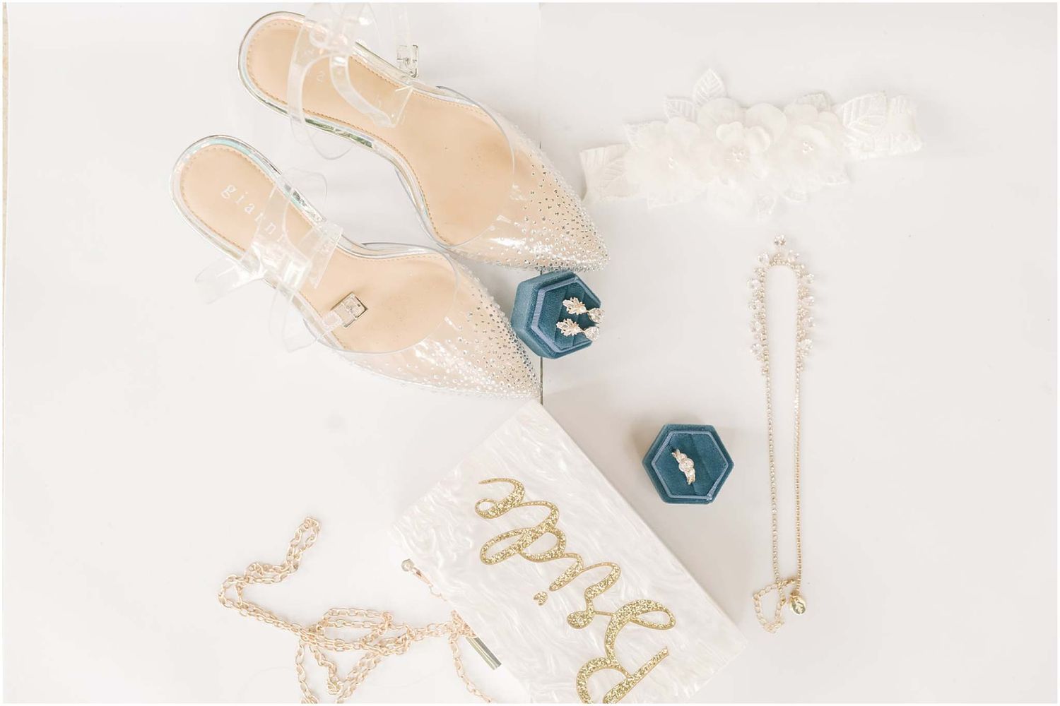Elegant bridal flat shoes, teal ring box, pearl necklace, and lace garter arranged on white surface.