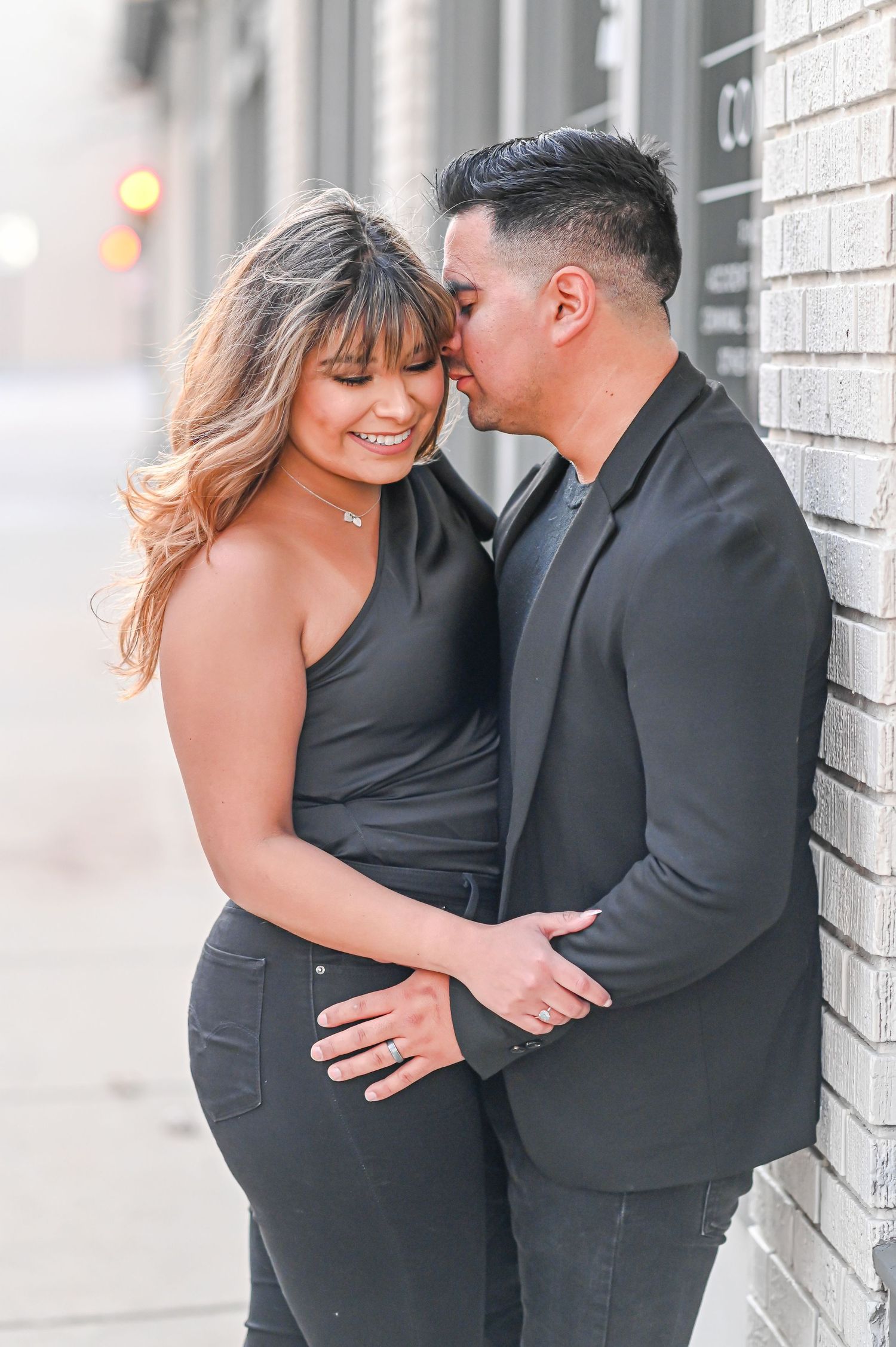 Man in black blazer kissing womans cheek while embracing against a white brick wall.