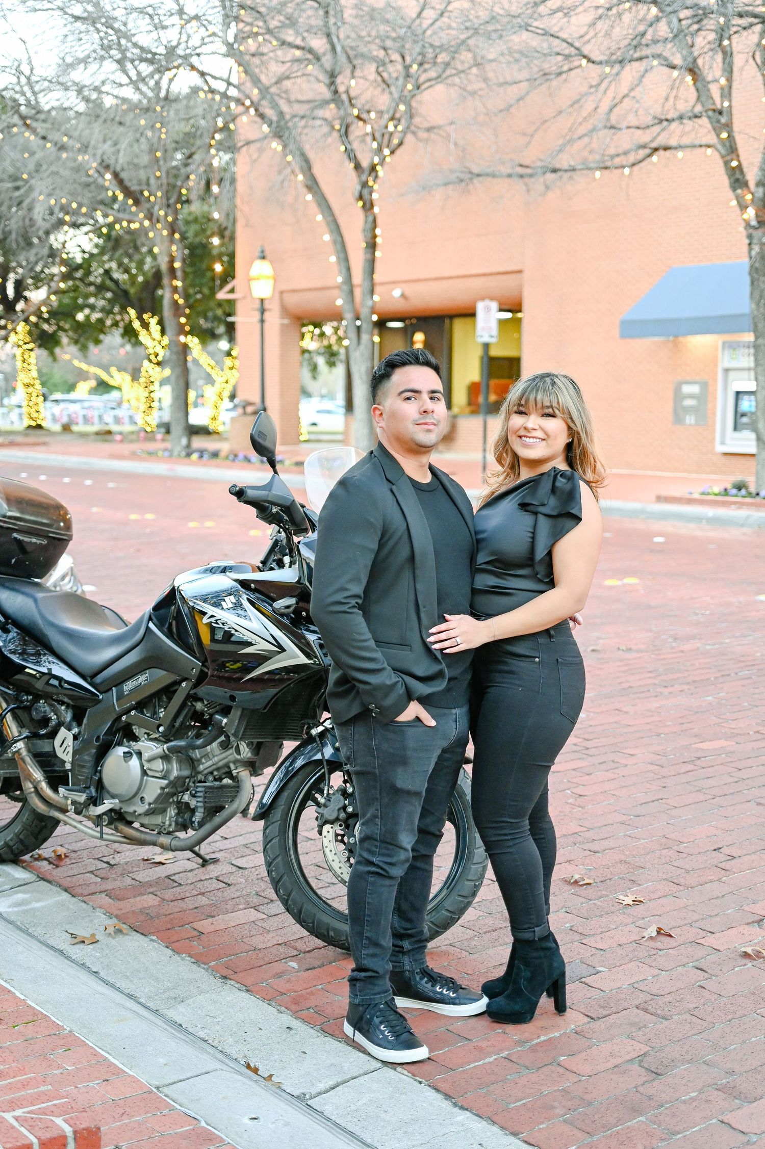 Couple in matching black outfits posing together near a motorcycle on a brick sidewalk in an urban setting.