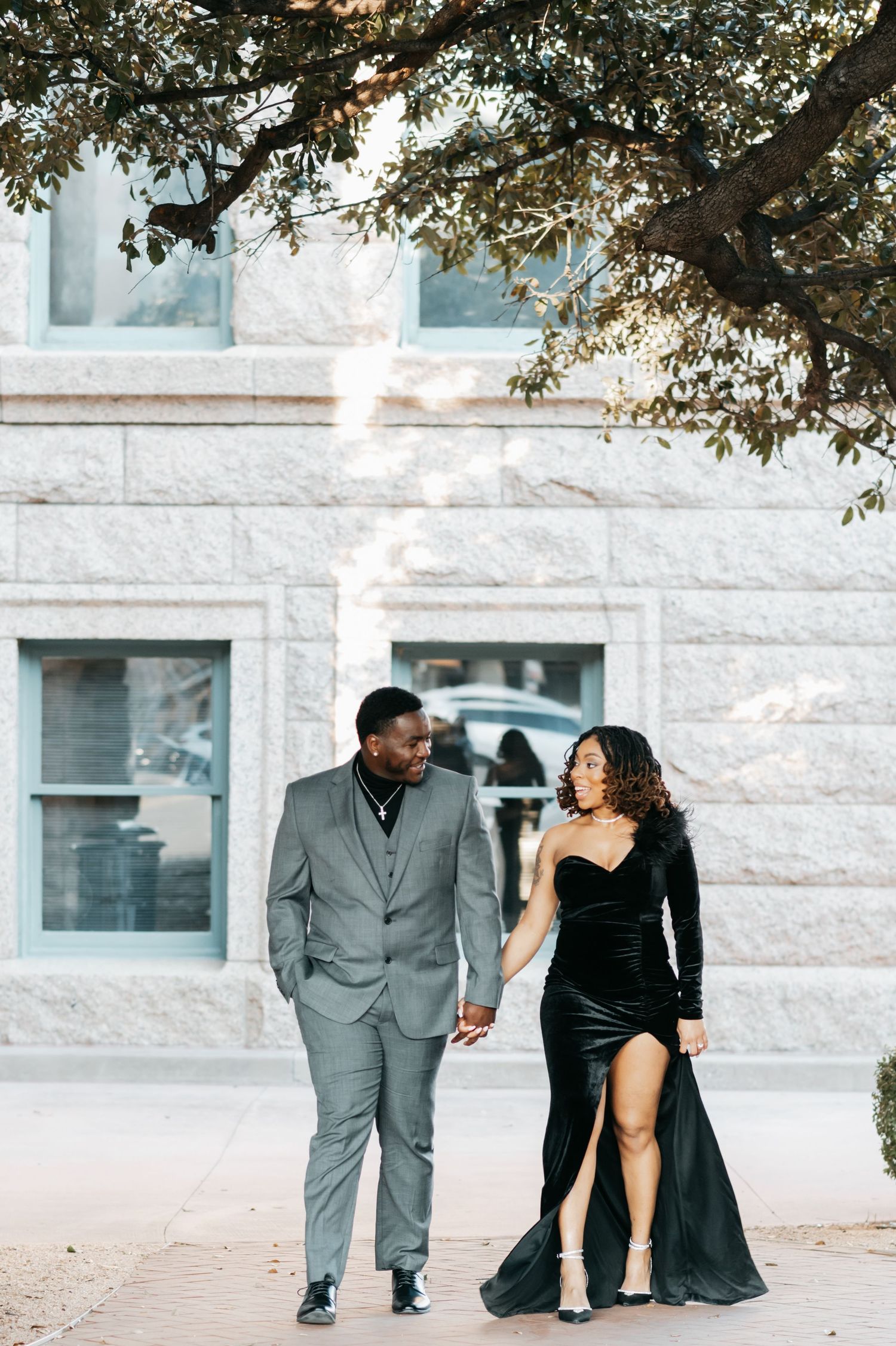 Couple holding hands walking in matching black velvet dress and gray suit outside a stone building.