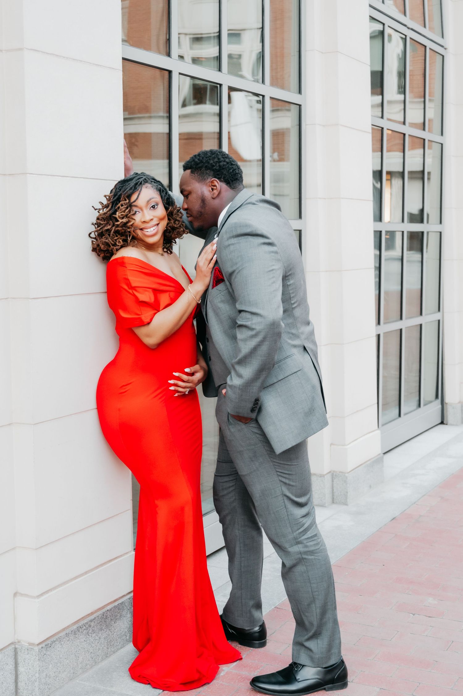 A couple poses romantically outside, woman in red gown smiles as man in gray suit leans in to kiss her cheek.