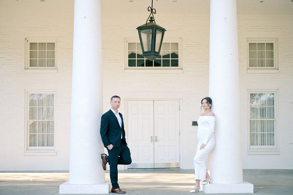 Elegant couple posing between white columns at a classic white building for an engagement photo session.