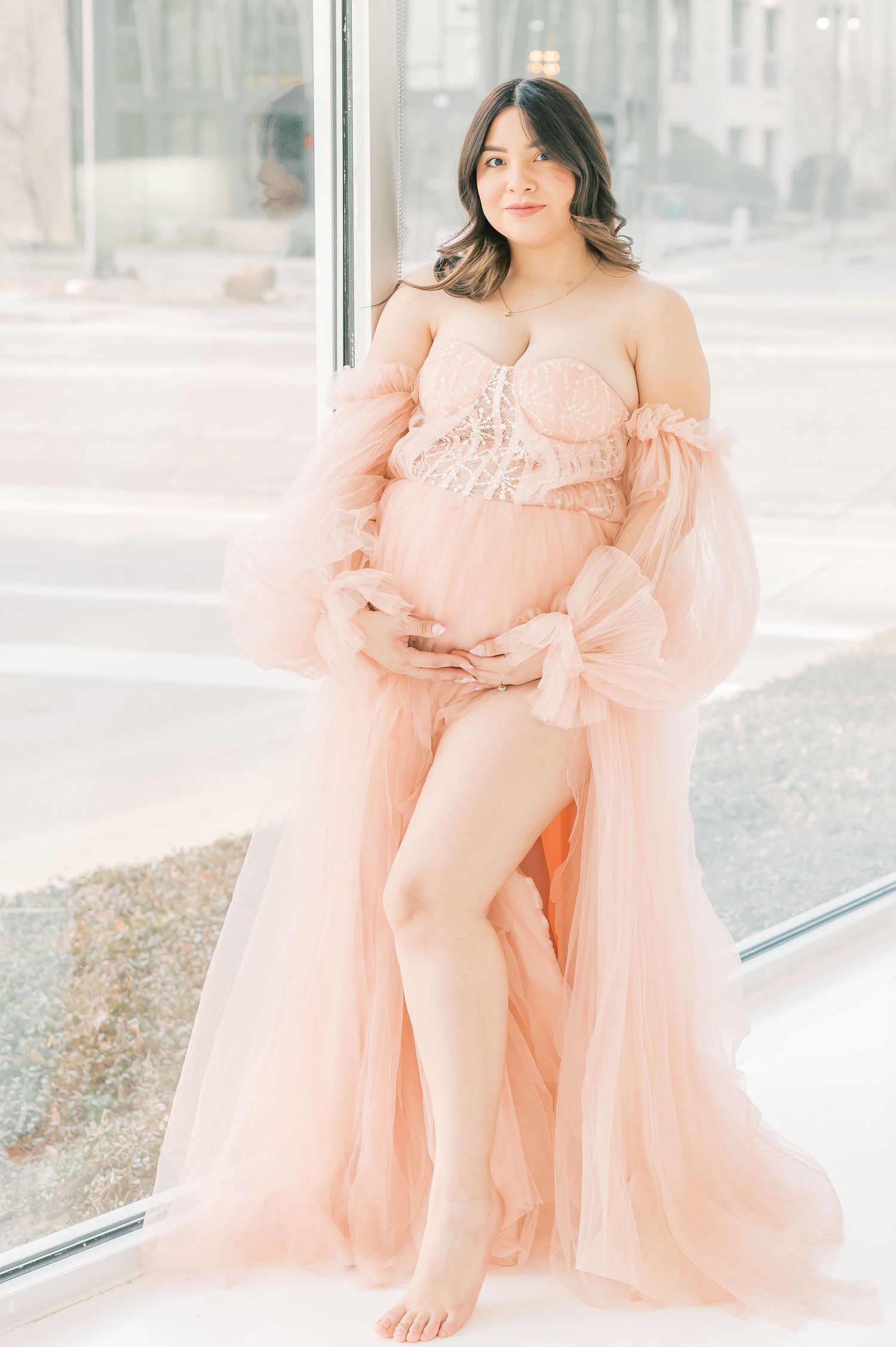Pregnant woman in a flowing pink tulle maternity gown posing by large windows in a bright studio.