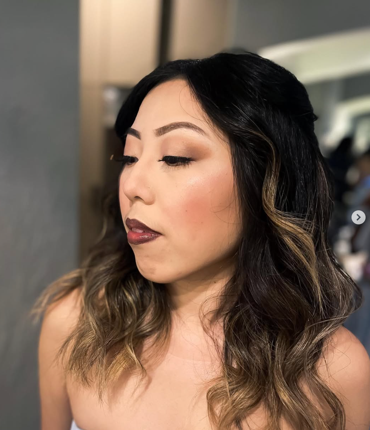Woman with dark balayage hair, glam makeup featuring rosy blush, defined brows, and a mauve lip, styled in soft waves.