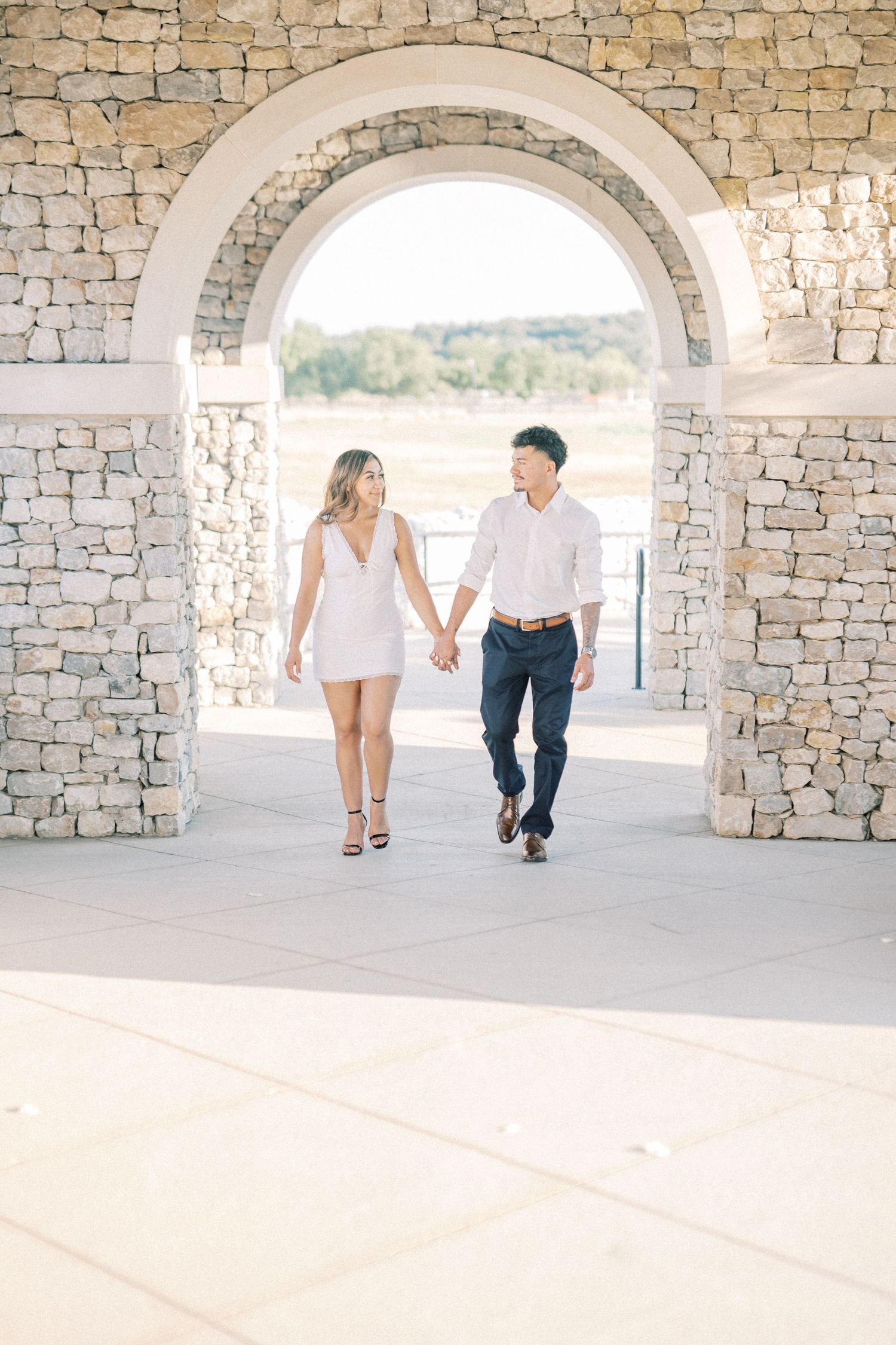 Couple holding hands walking through stone archway during outdoor engagement session.