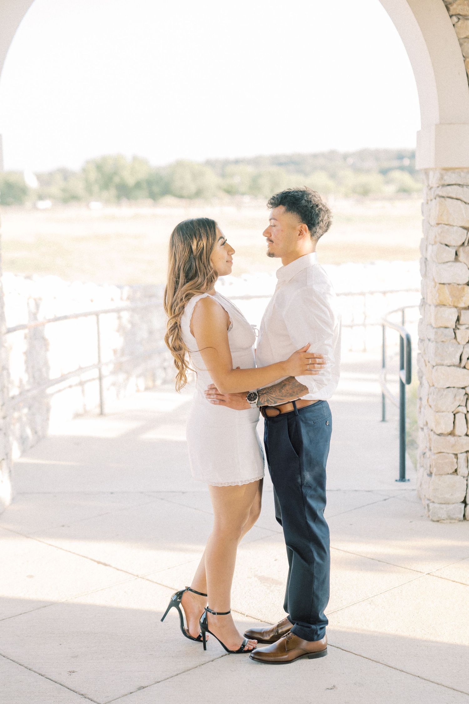 A couple in elegant attire embraces and gazes at each other under an arch during an outdoor engagement session.
