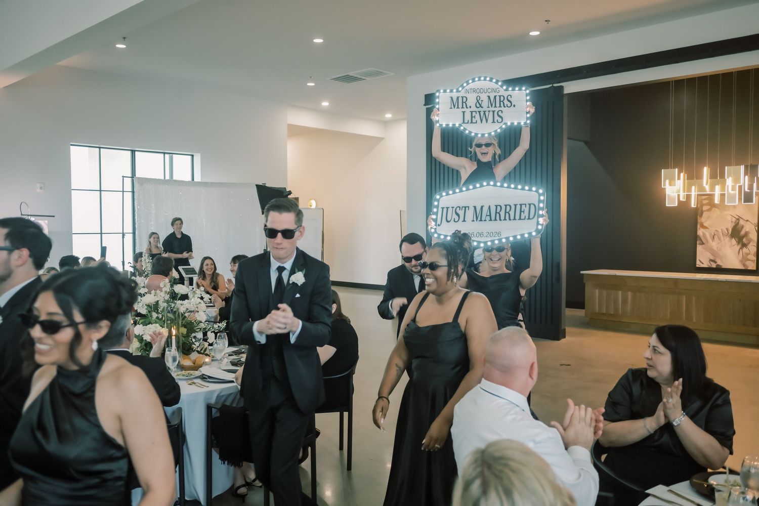 Wedding guests in black attire celebrate as a couple enters with a glowing Mr and Mrs Just Married marquee sign.