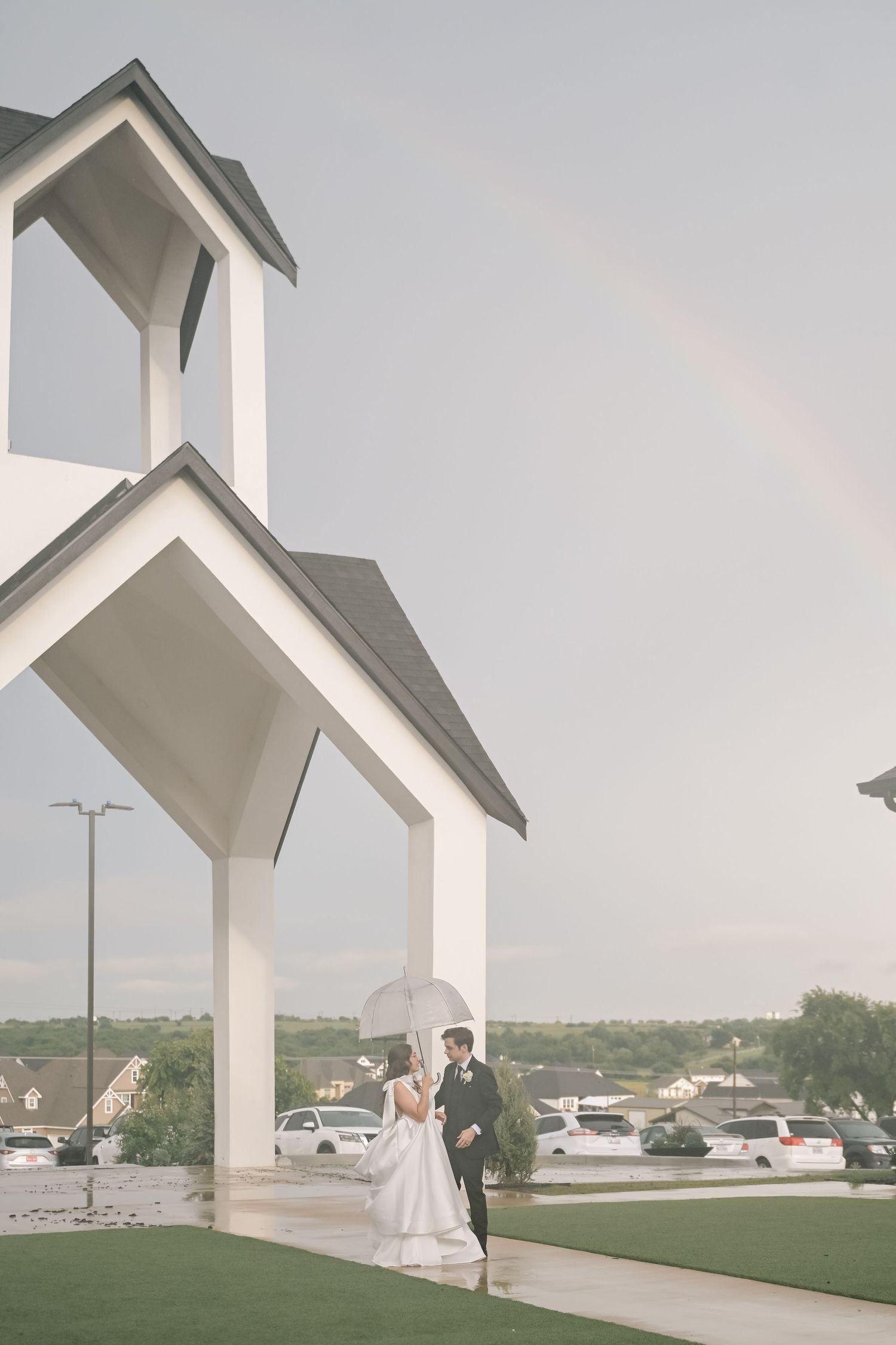 Wedding couple with umbrella pose near white chapel entrance with rainbow in rainy sky.