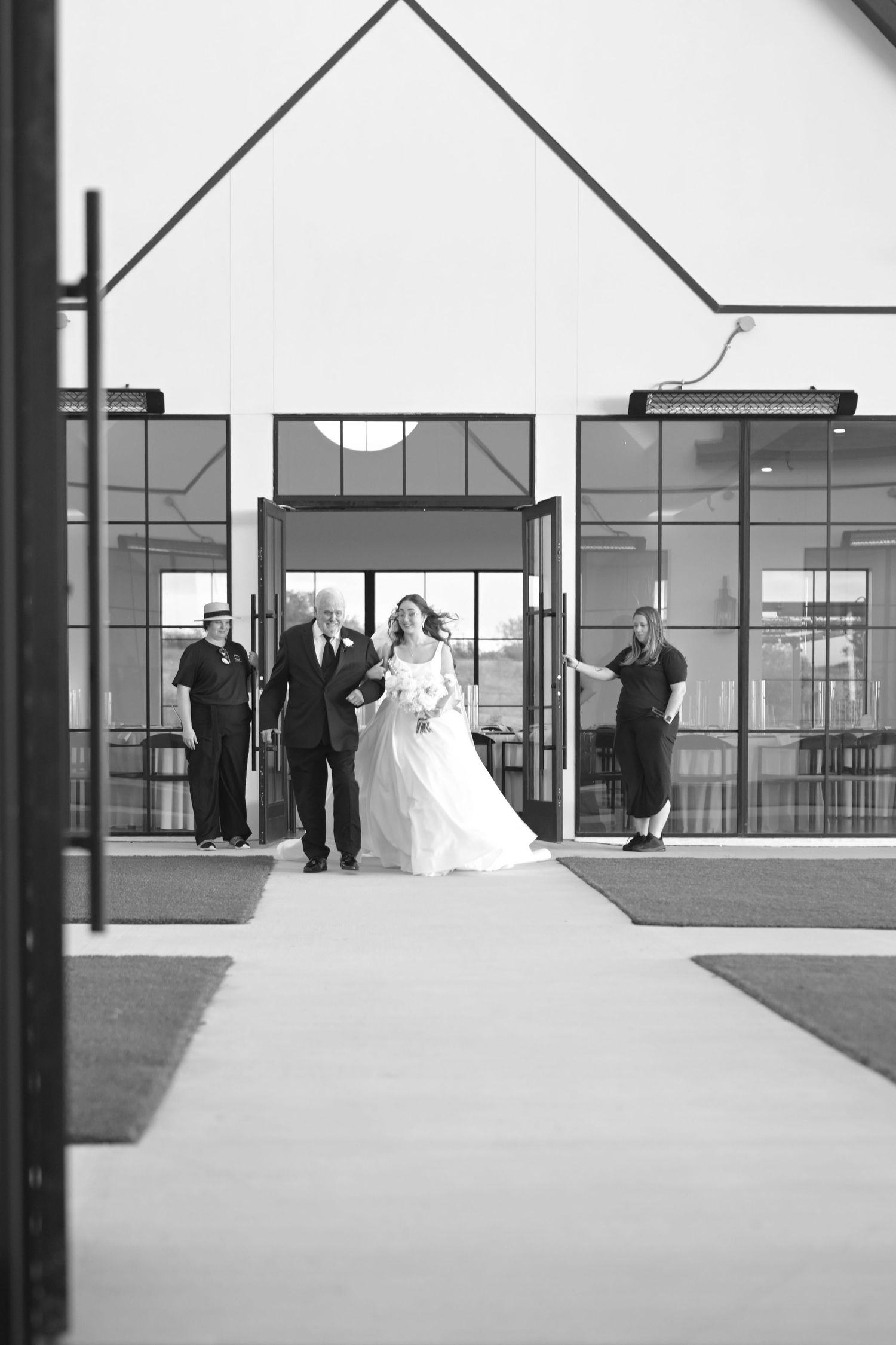 Bride walks down the aisle with escort outside a modern white chapel during an elegant wedding ceremony.