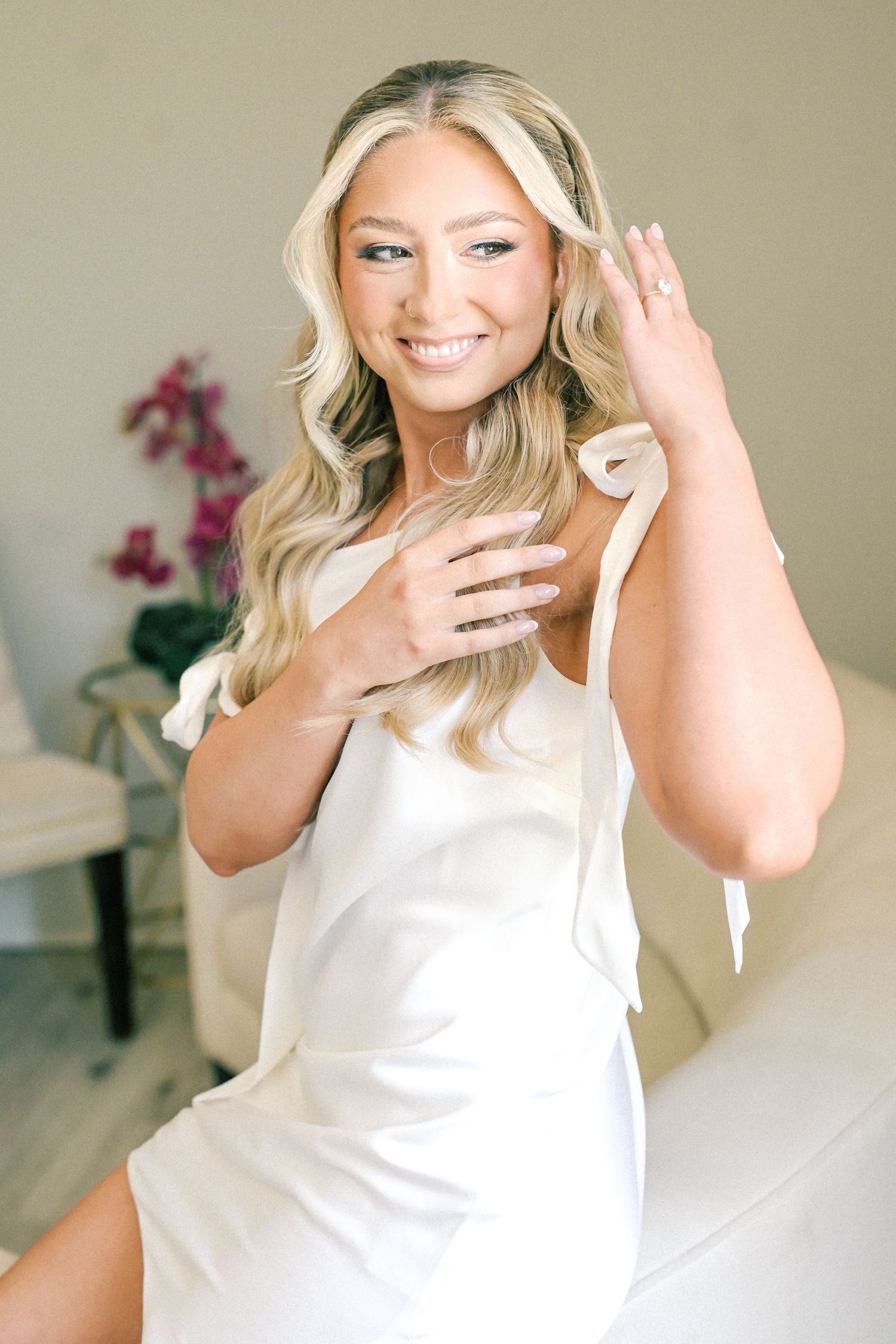 Smiling blonde bride in white slip dress shows off engagement ring while seated before wedding ceremony.