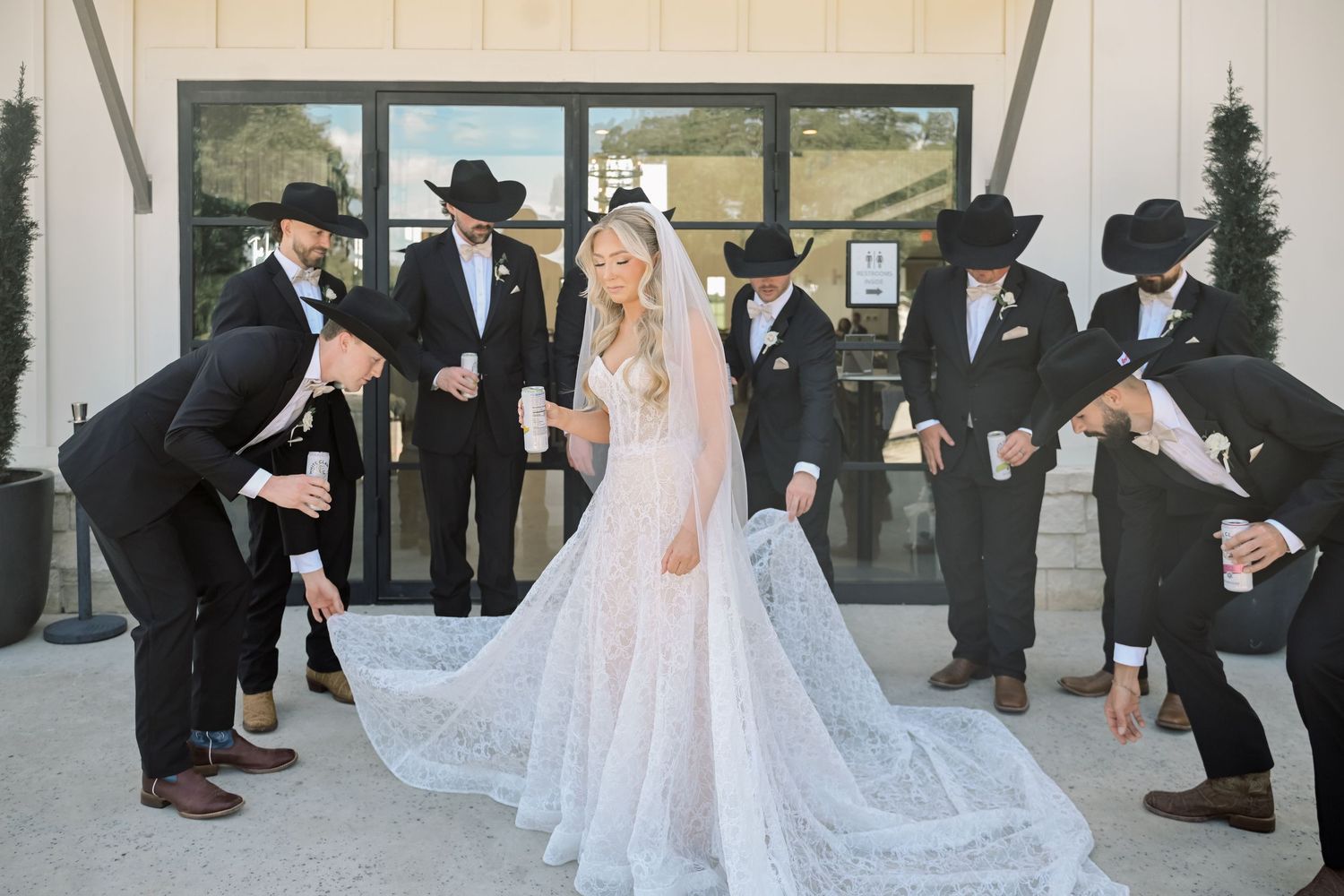 Bride in lace gown surrounded by groomsmen in black suits and cowboy hats outside wedding venue.