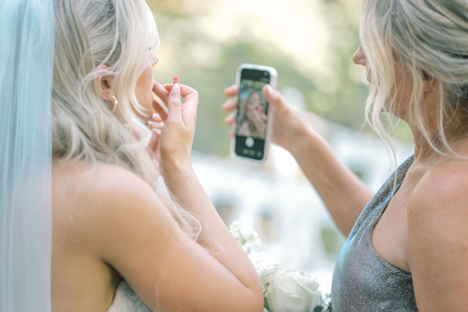 Bridesmaid in silver dress photographs bride adjusting earring with smartphone outdoors.