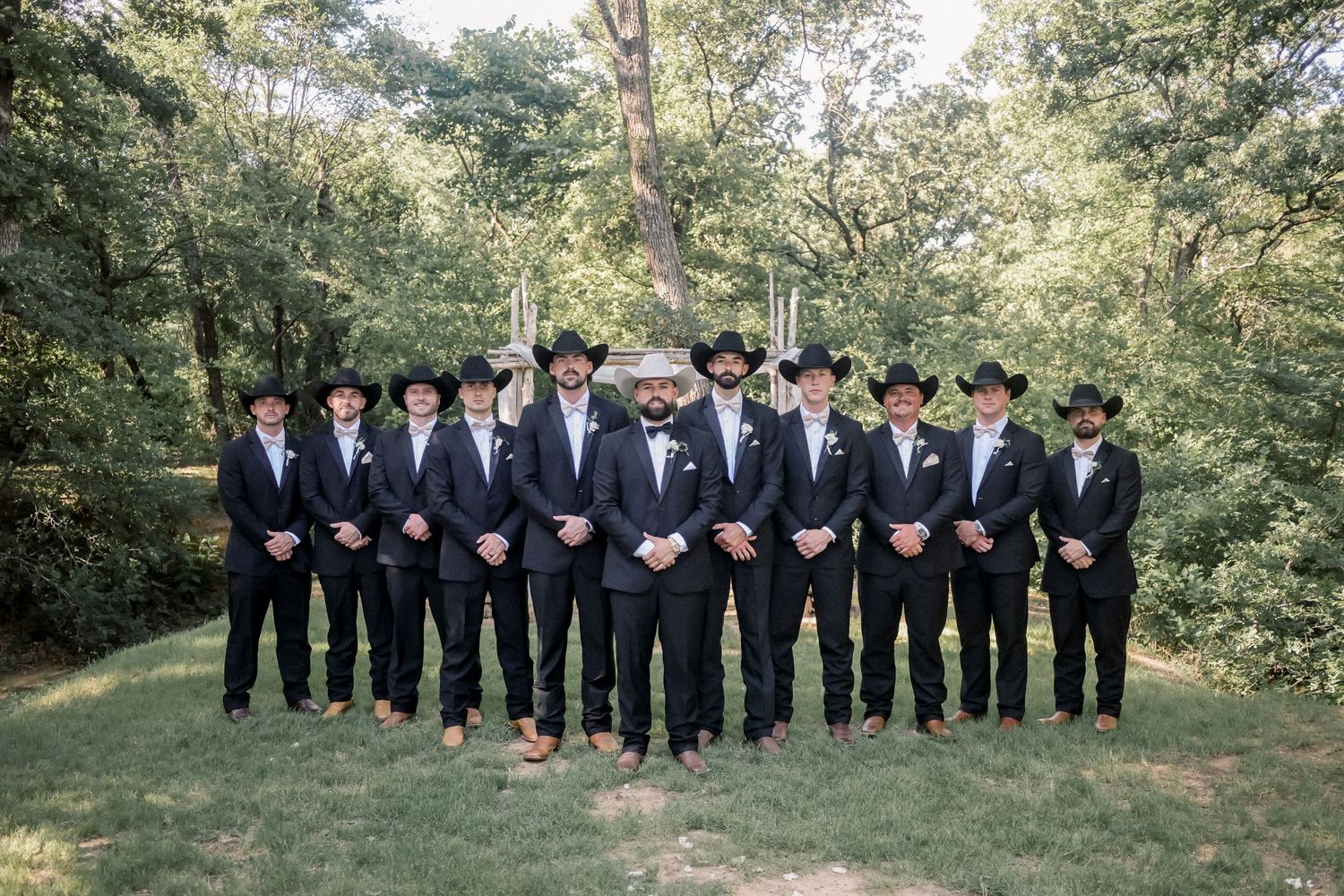 Groom and groomsmen in black suits and cowboy hats pose outdoors at a rustic country wedding.