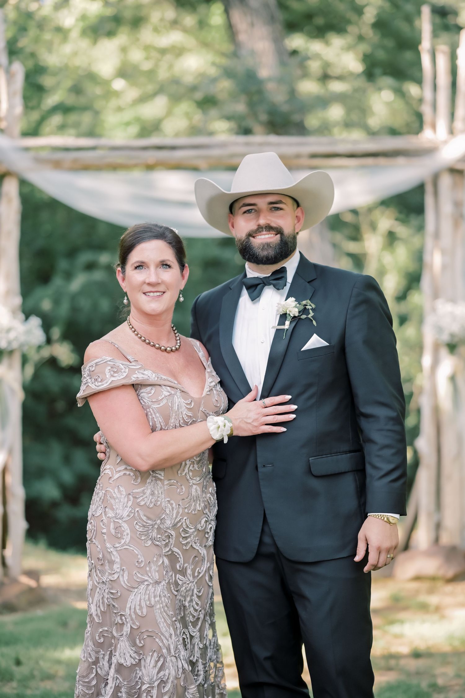 Groom in black tuxedo and white cowboy hat posing with woman in gold lace gown at outdoor wedding.