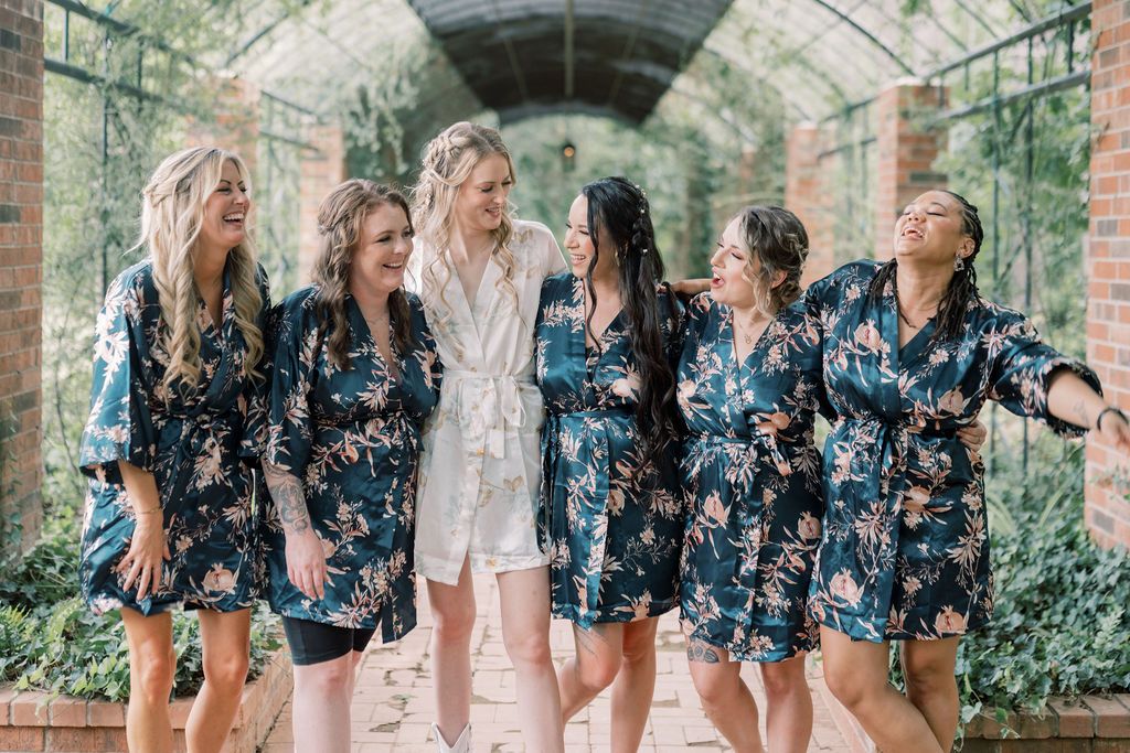 Six bridesmaids in navy floral robes laugh together outdoors, with the bride in white at center.