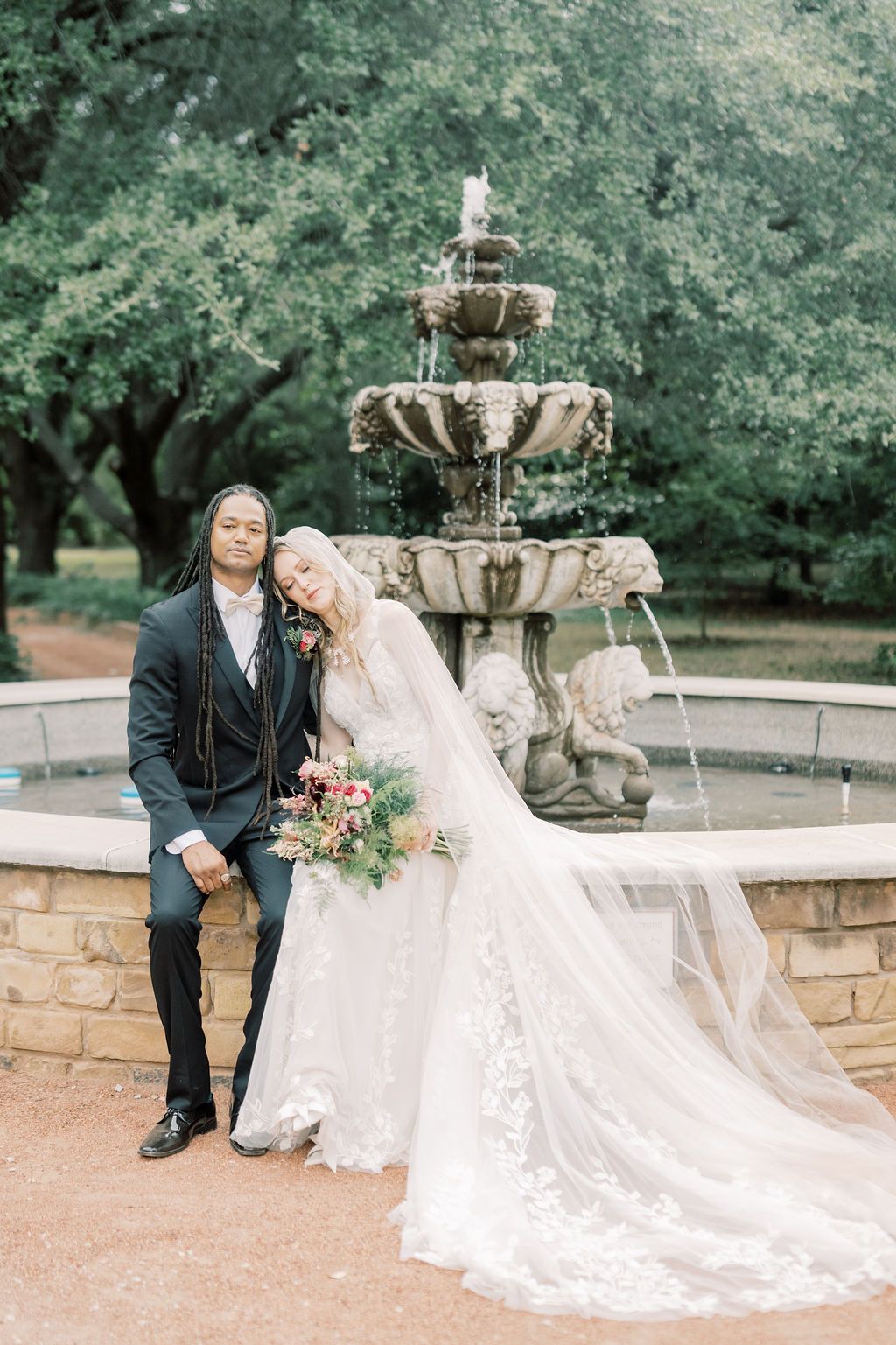 Bride and groom pose by ornate stone fountain in lush garden, bride in lace gown with flowing veil holding bouquet.