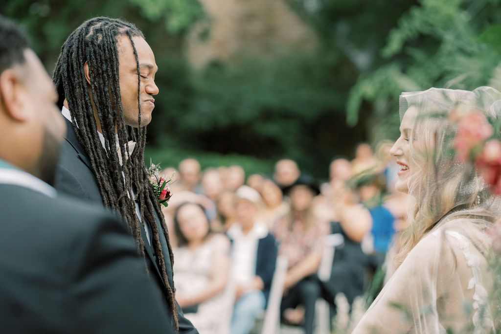 Groom with long dreadlocks smiling at bride during outdoor wedding ceremony with guests seated in background.