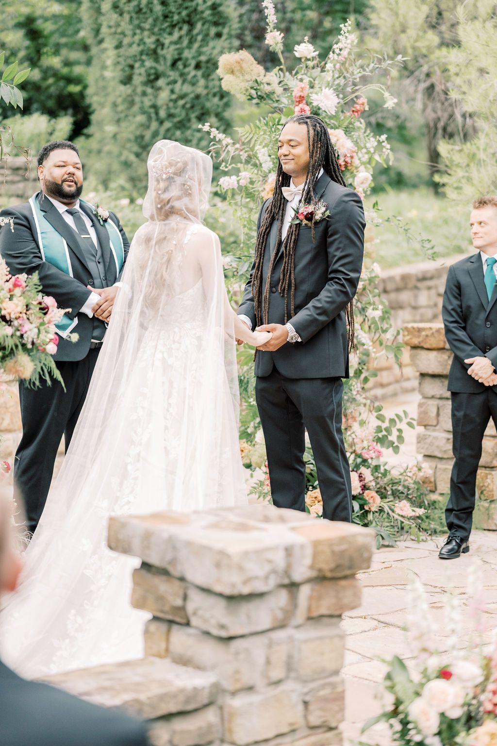 Bride and groom holding hands at outdoor wedding ceremony with floral arch and groomsmen standing nearby.